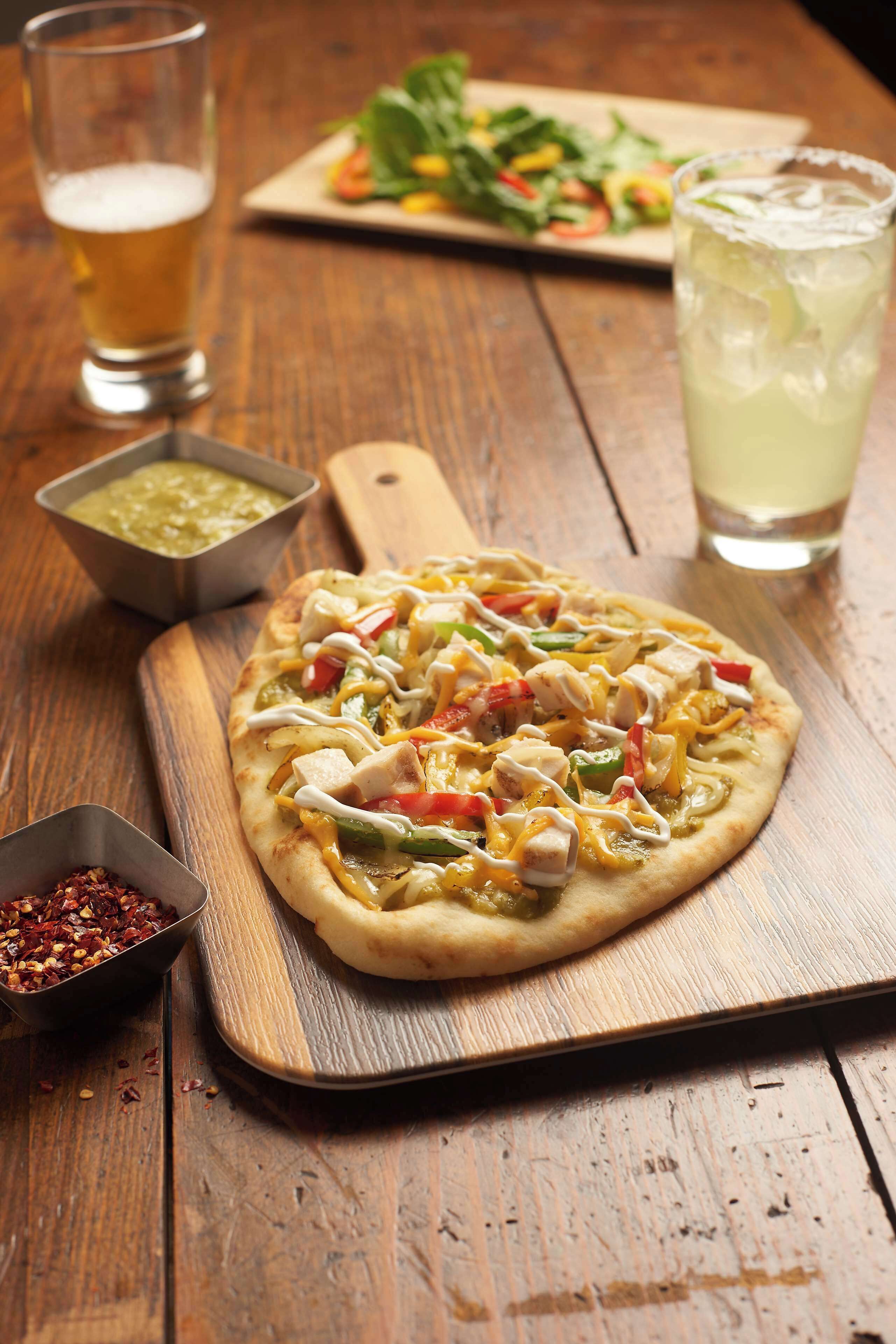 Une pizza garnie de divers ingrédients, servie sur une planche en bois, accompagnée d’une salade, d’une boisson et d’un petit bol de sauce.
