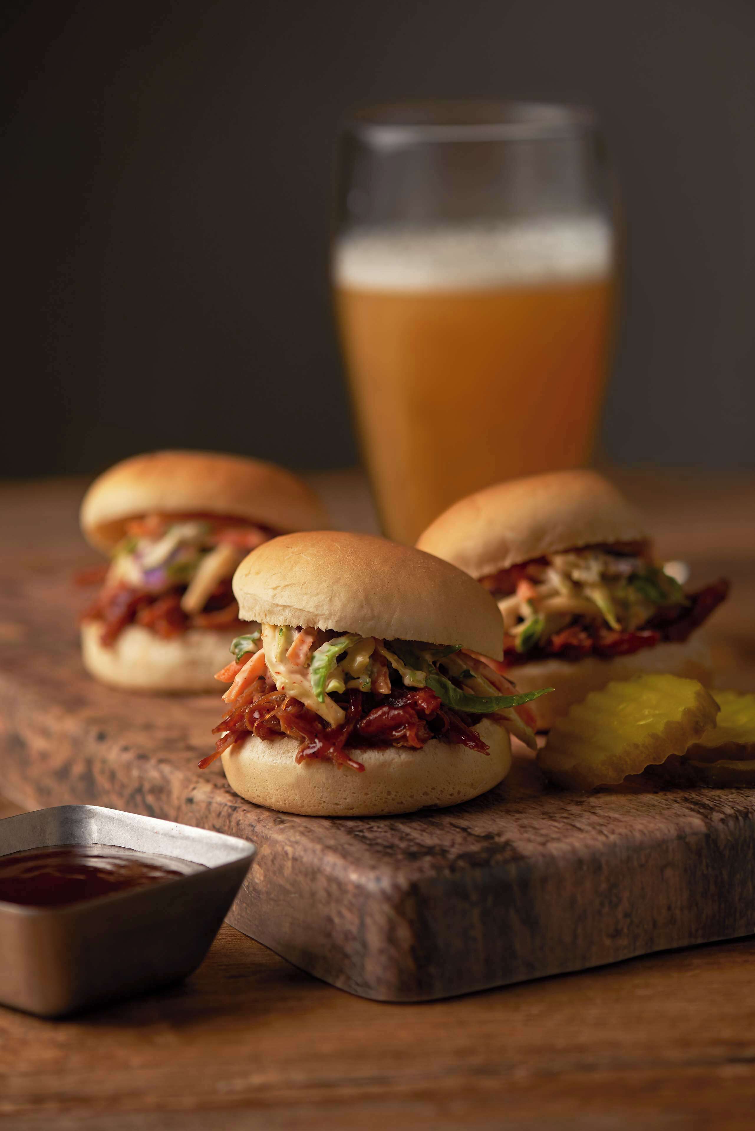 Two pulled pork sandwiches on buns, a glass of beer, and a small bowl of sauce on a wooden board.