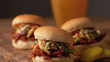 Two pulled pork sandwiches on buns, a glass of beer, and a small bowl of sauce on a wooden board.