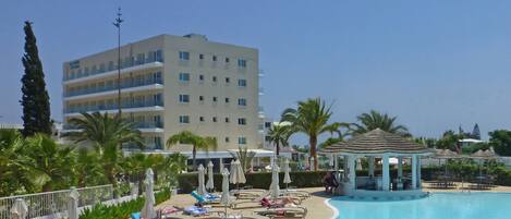 A pool area with lounge chairs and umbrellas, a gazebo, and a multi-story building in the background.