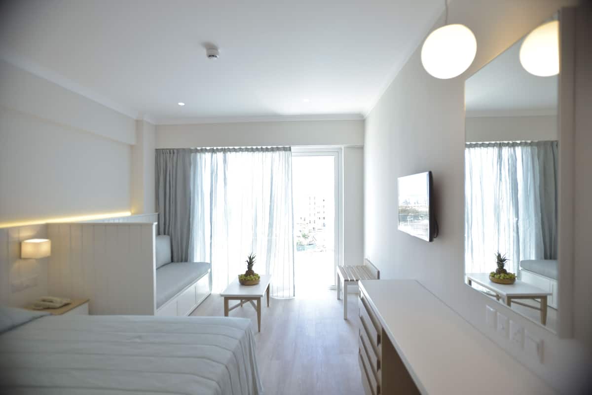 A hotel room with a bed, a desk, a chair, a small table with fruit, a mirror, and a window with curtains.