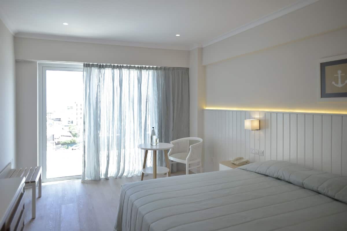 A hotel room with a bed, a small table with a chair, a bedside telephone, and a view through a large window with curtains.