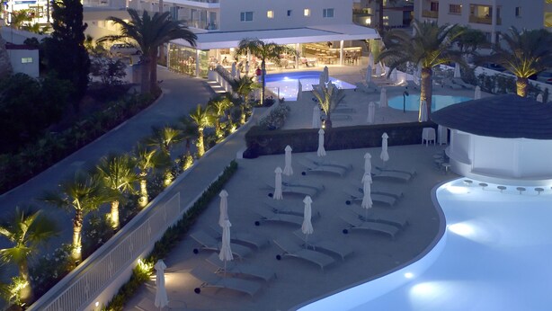 Zona de piscina hotelera con tumbonas y sombrillas, rodeada de palmeras y un edificio blanco al fondo.