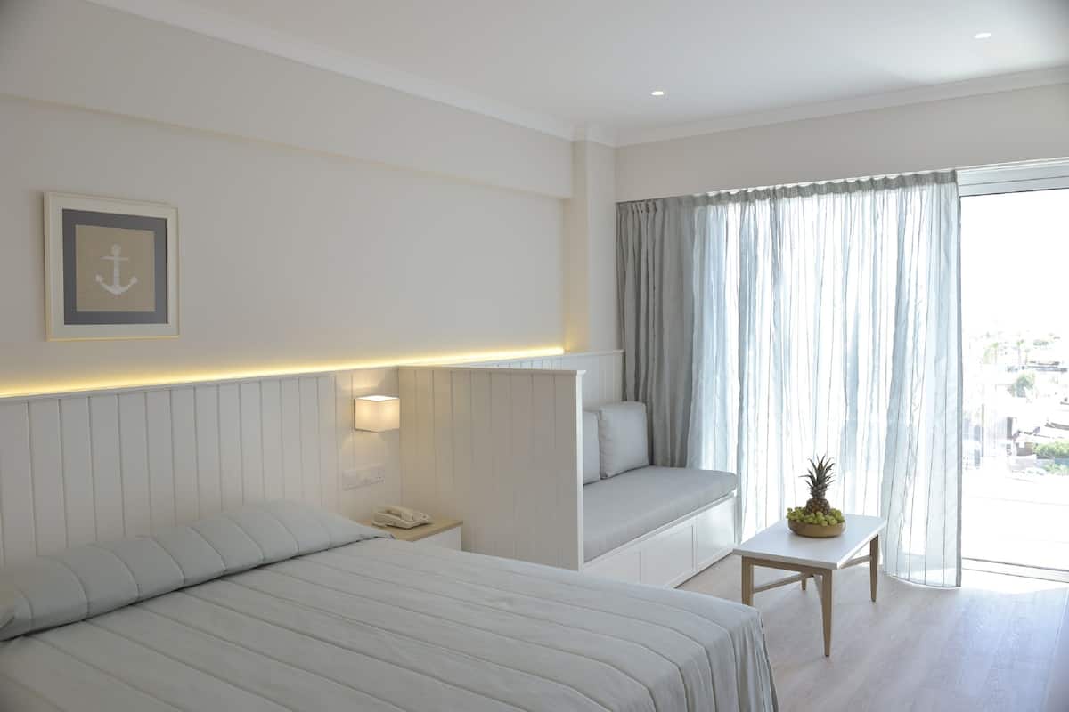 A hotel room with a bed, a bedside table with a phone, a small table with fruit, a sofa, and a window with curtains.