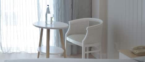 Habitación de hotel con cama, una silla blanca, una mesita redonda con una botella y un vaso, y un teléfono sobre una mesita de noche.