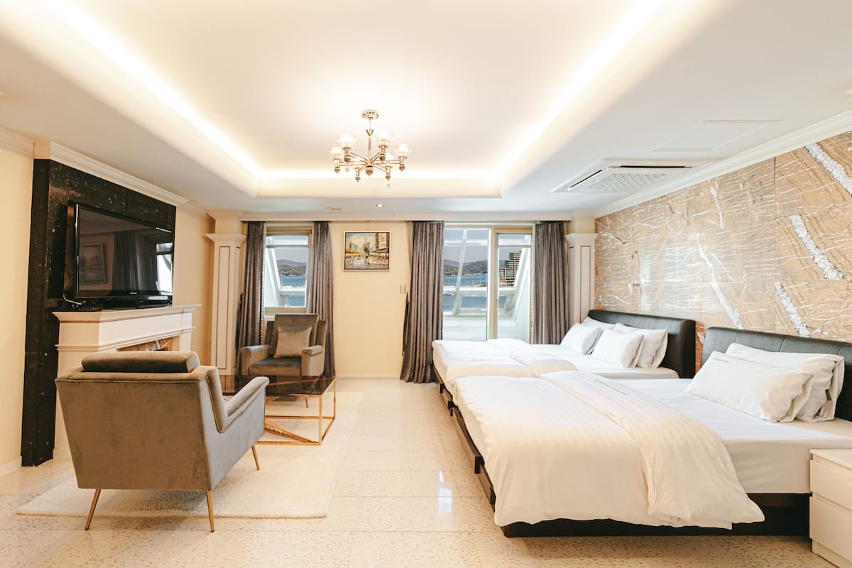 A hotel room with a large bed, a desk with a flat-screen TV, a chair, and a view of the sea through the windows.