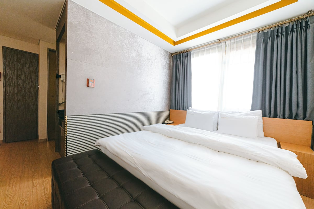 A modern hotel room with a large bed, a wooden bench, a bedside table, and a window with curtains.