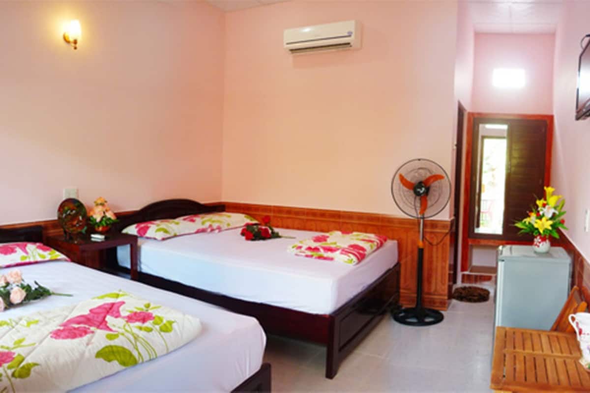 Habitación de hotel con dos camas, una mesita de noche con lámpara, un ventilador, un refrigerador pequeño y una silla de madera.