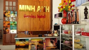 A reception area with a wooden sign reading "MINH ANH Garden hotel," a desk with various items, a decorative vase, and a shelf with boxes. - Minh Anh Garden Hotel (Mui Ne)