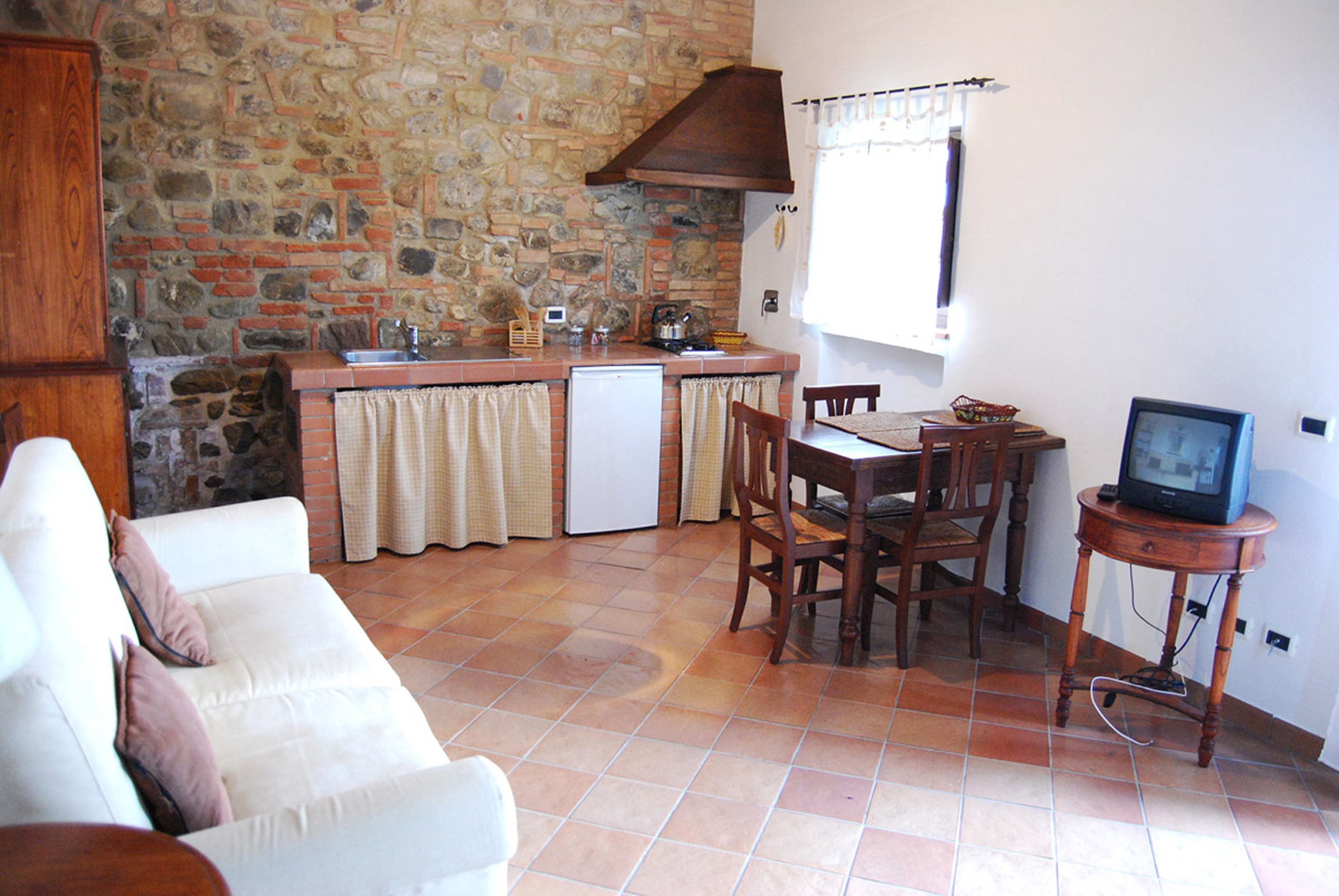 Una cocina compacta con pared de piedra, un refrigerador blanco, una mesa de comedor de madera con sillas y un televisor sobre un mueble de madera.