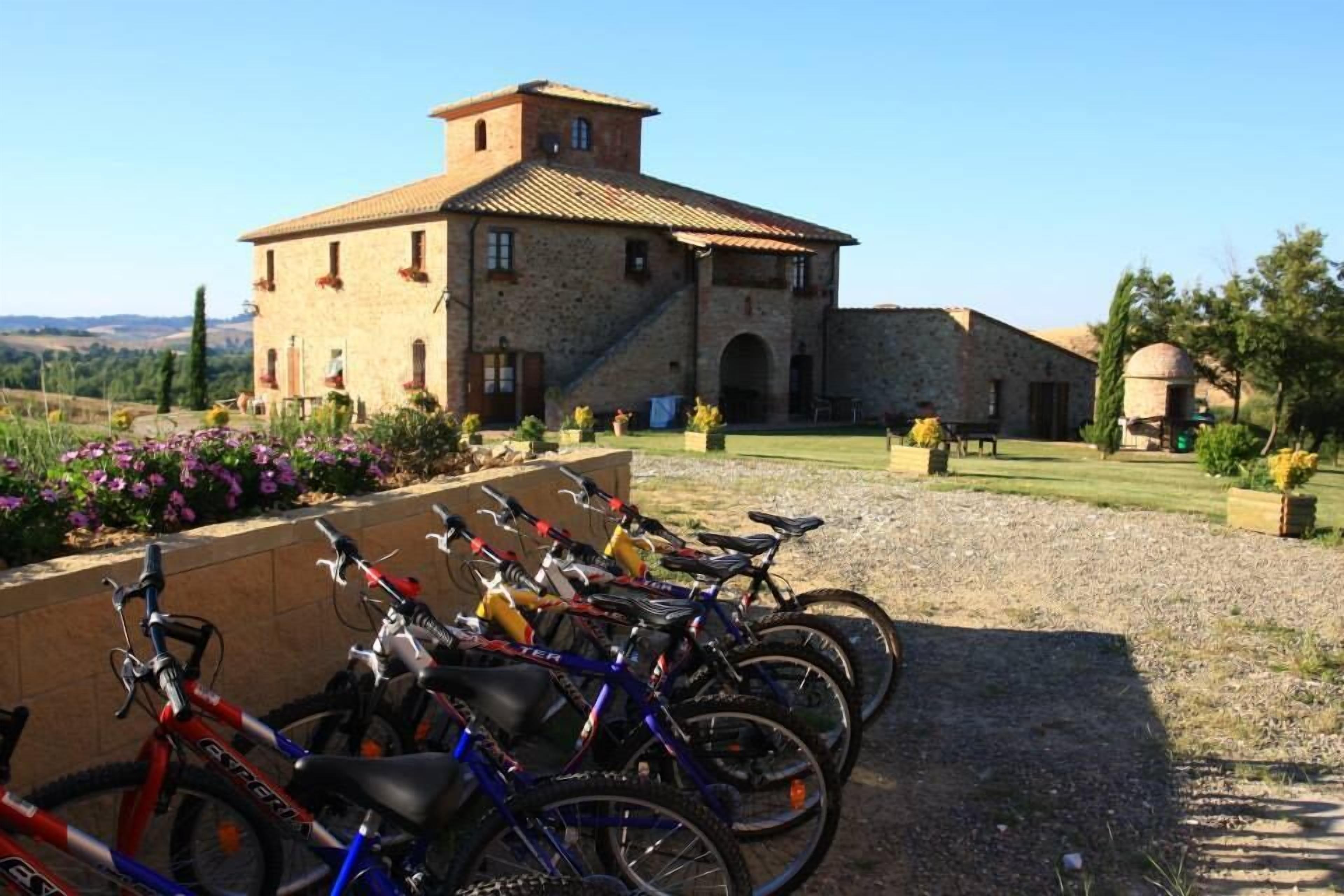 Une rangée de vélos est garée devant un bâtiment en pierre, doté d’un toit en tuiles, et entouré d’un chemin de gravier et de parterres de fleurs.