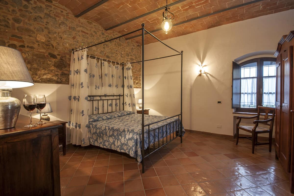 A four-poster bed with a floral bedspread, a wooden nightstand with a lamp, and a chair by a window with curtains.