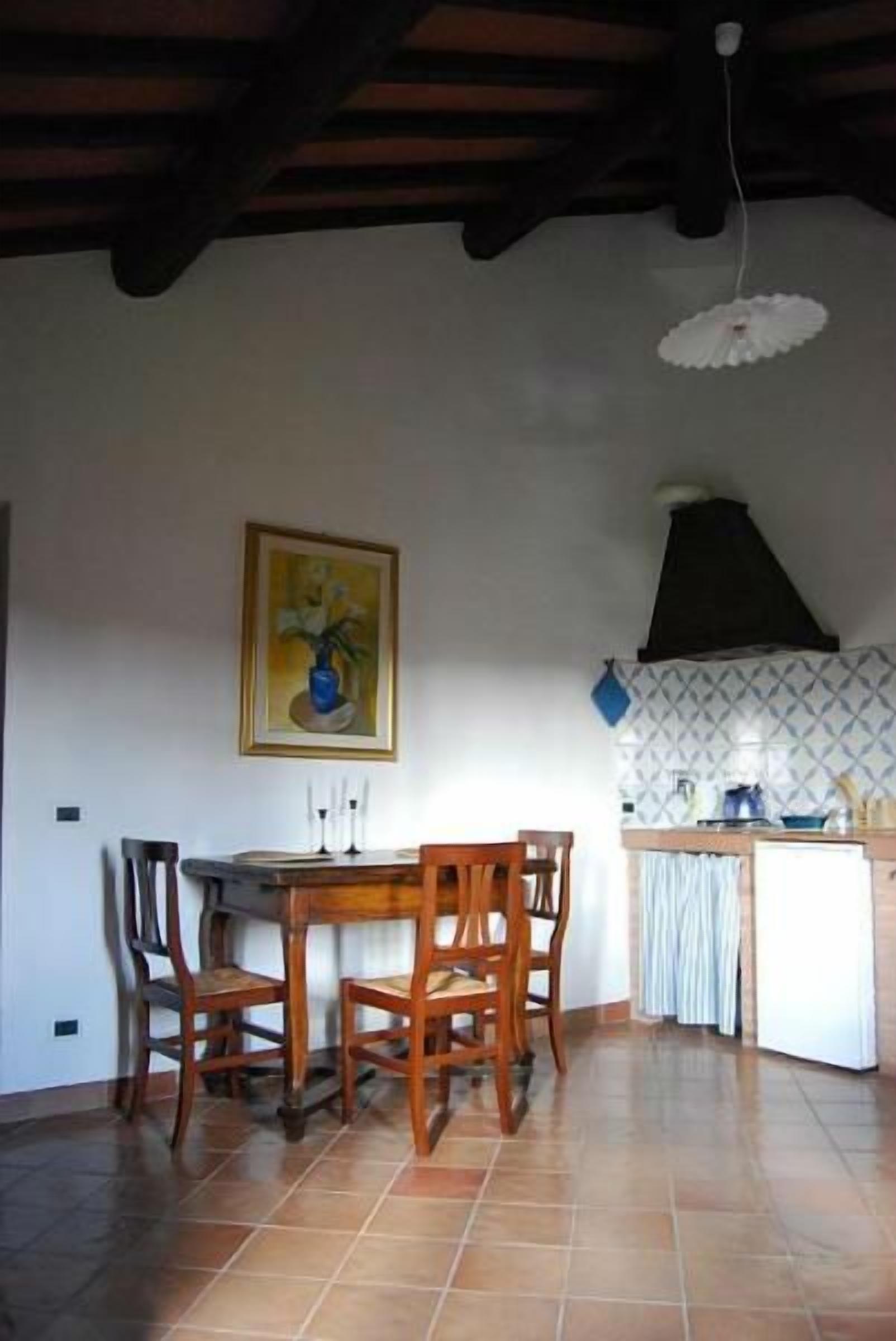 A dining area with a wooden table and chairs, a painting on the wall, and a kitchen area with a stove and cabinets.
