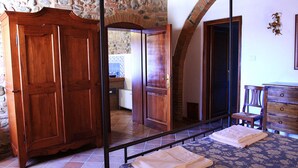 A bedroom with a large mirror reflecting a bathroom, a wooden wardrobe, a bed with a patterned bedspread, and a small wooden chair. - Agriturismo Ragoncino (Lajatico)