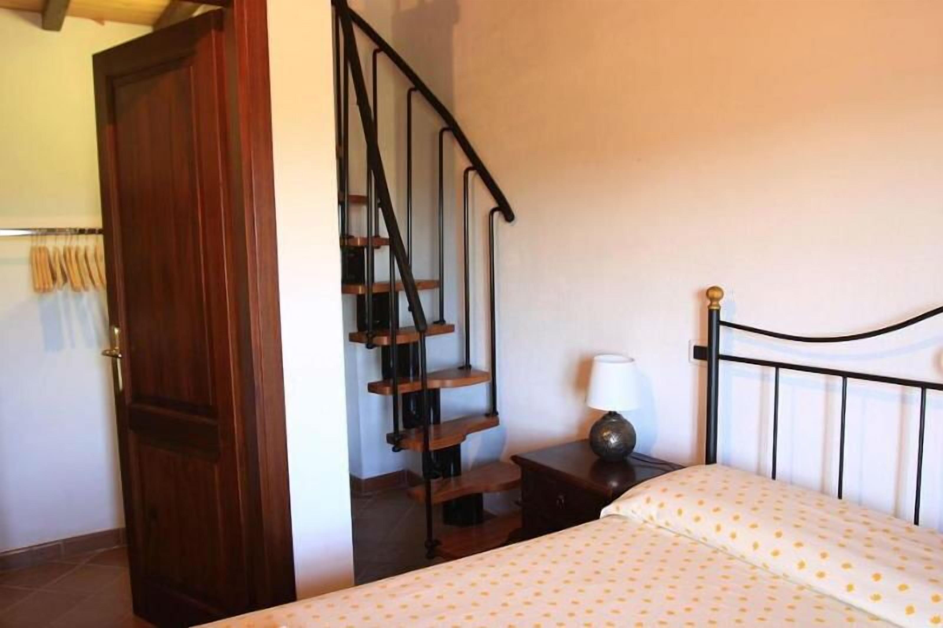 A bedroom with a wooden door, a staircase, a bed with a white and yellow polka dot bedspread, and a bedside table with a lamp.