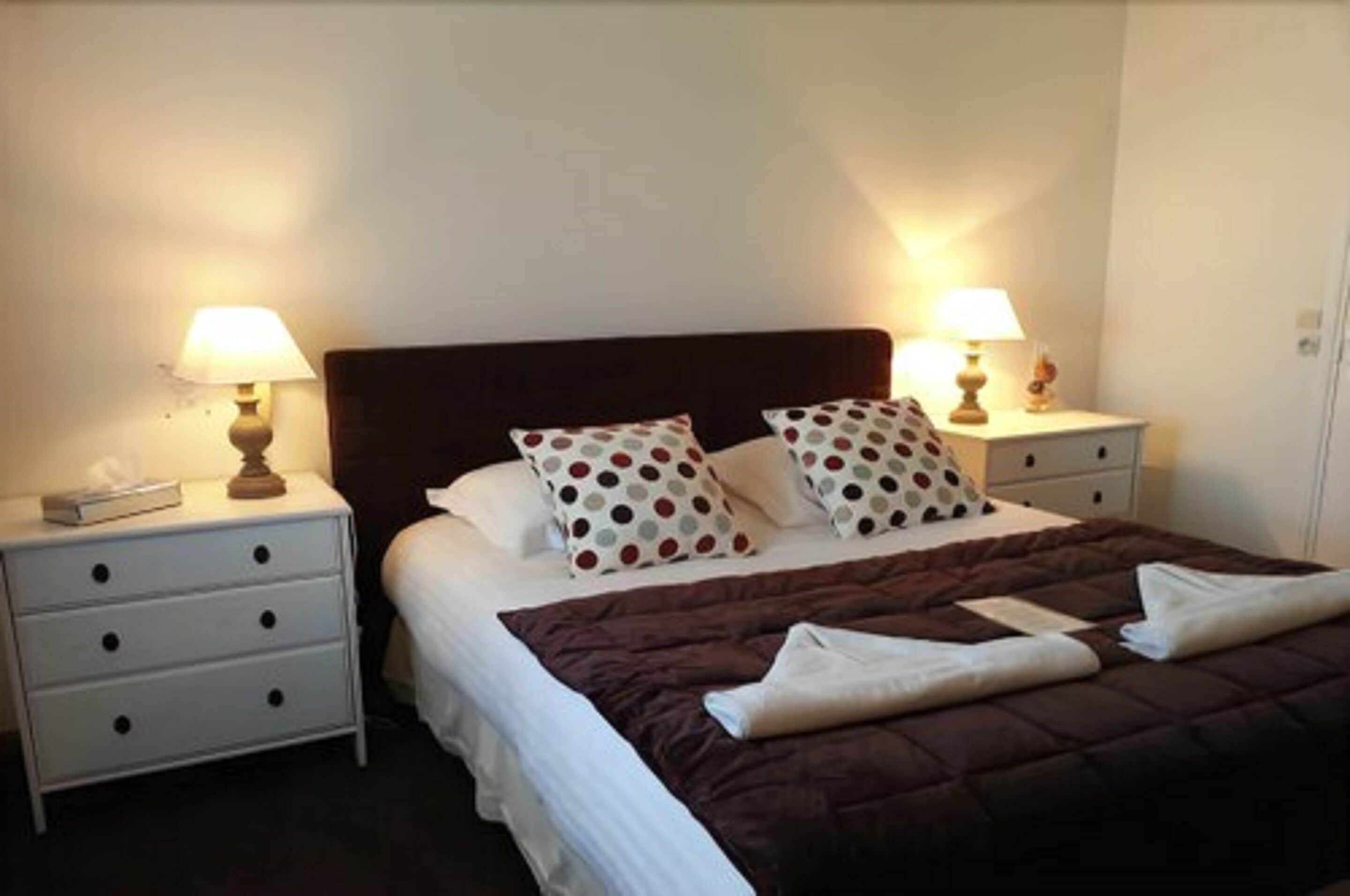 A neatly made bed with a brown quilt, white pillows with polka dots, and two bedside lamps on white nightstands.