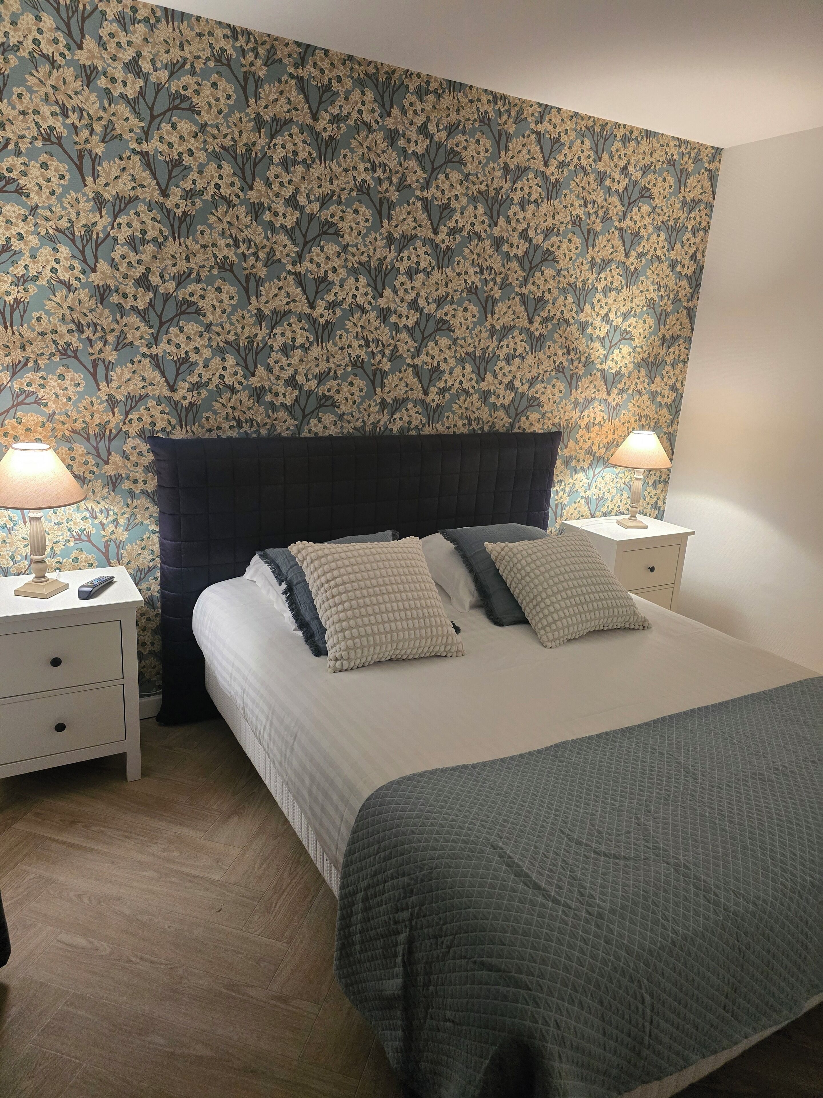 A bedroom with a floral wallpaper, a dark headboard, two bedside tables with lamps, and a bed with a quilted cover.
