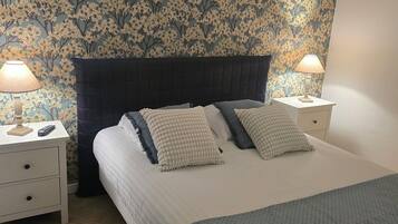 A bedroom with a floral wallpaper, a dark headboard, two bedside tables with lamps, and a bed with a quilted cover.