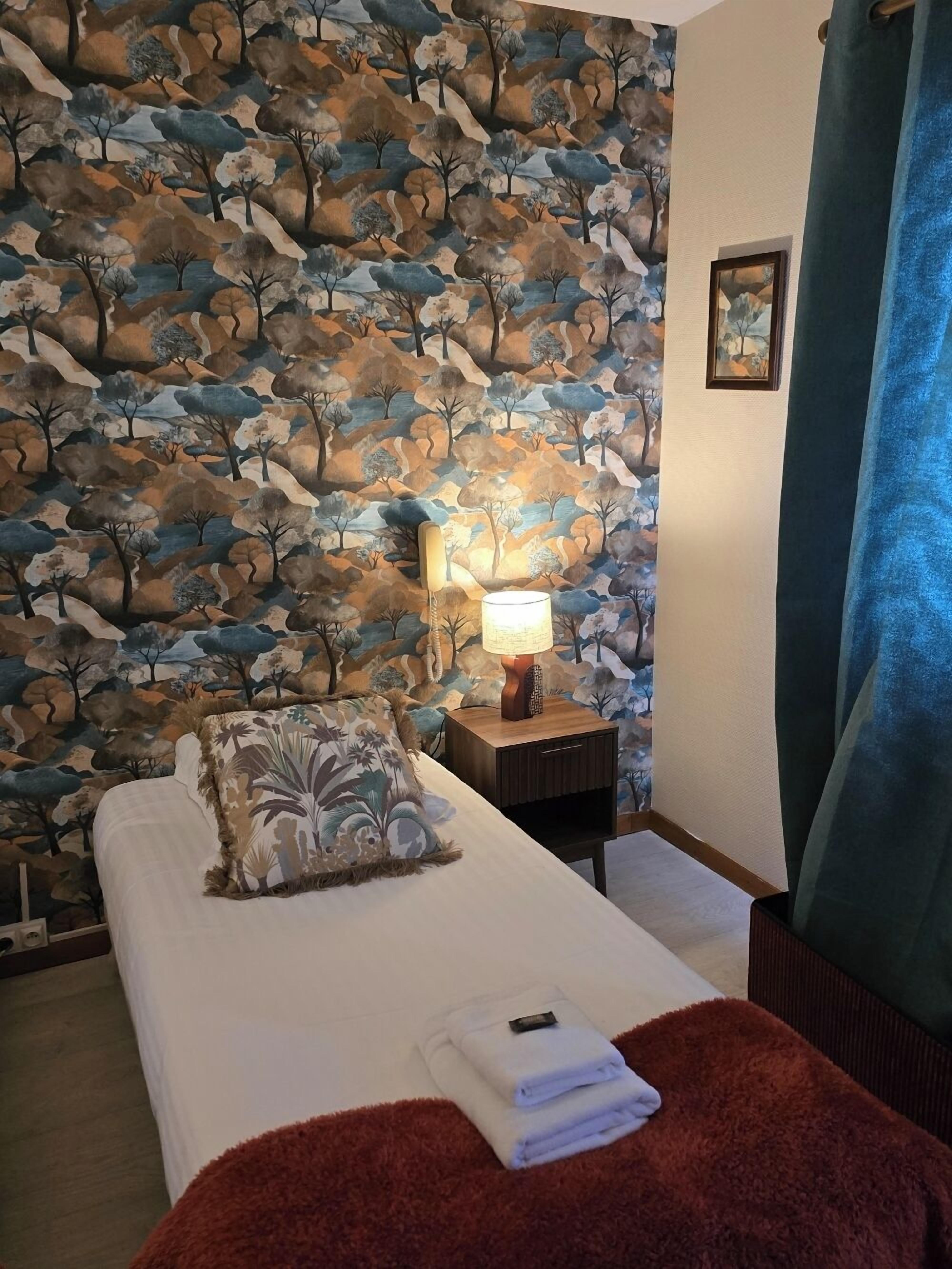 Une chambre d’hôtel avec un lit, une table de chevet avec une lampe, un tableau encadré au mur et un rideau bleu.