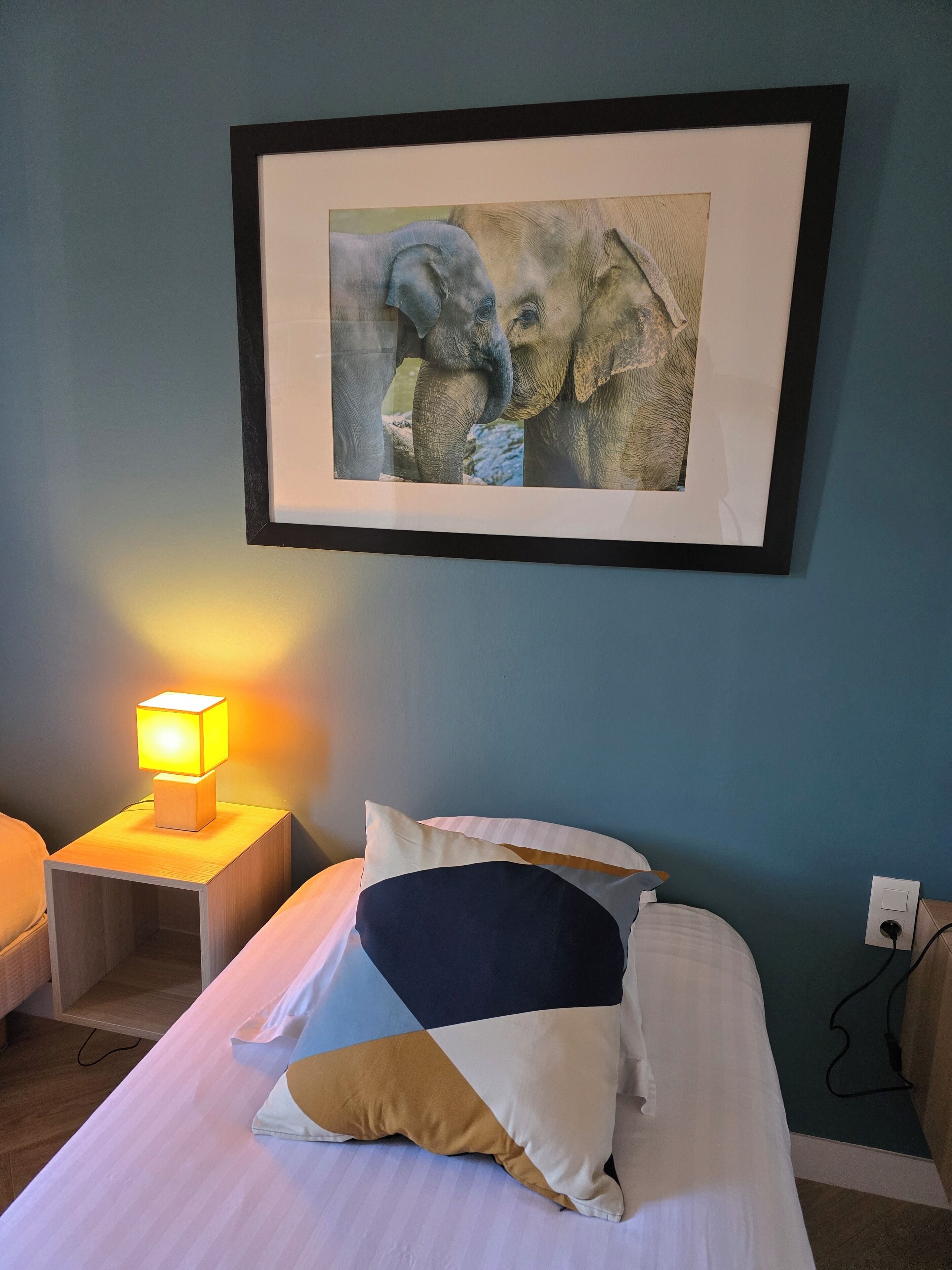 A bedroom with a bed, pillows, a nightstand with a lamp, and a framed picture on the wall.