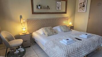 Quarto bem arrumado com cama, mesas de cabeceira, uma cadeira e uma pequena mesa com chávenas.