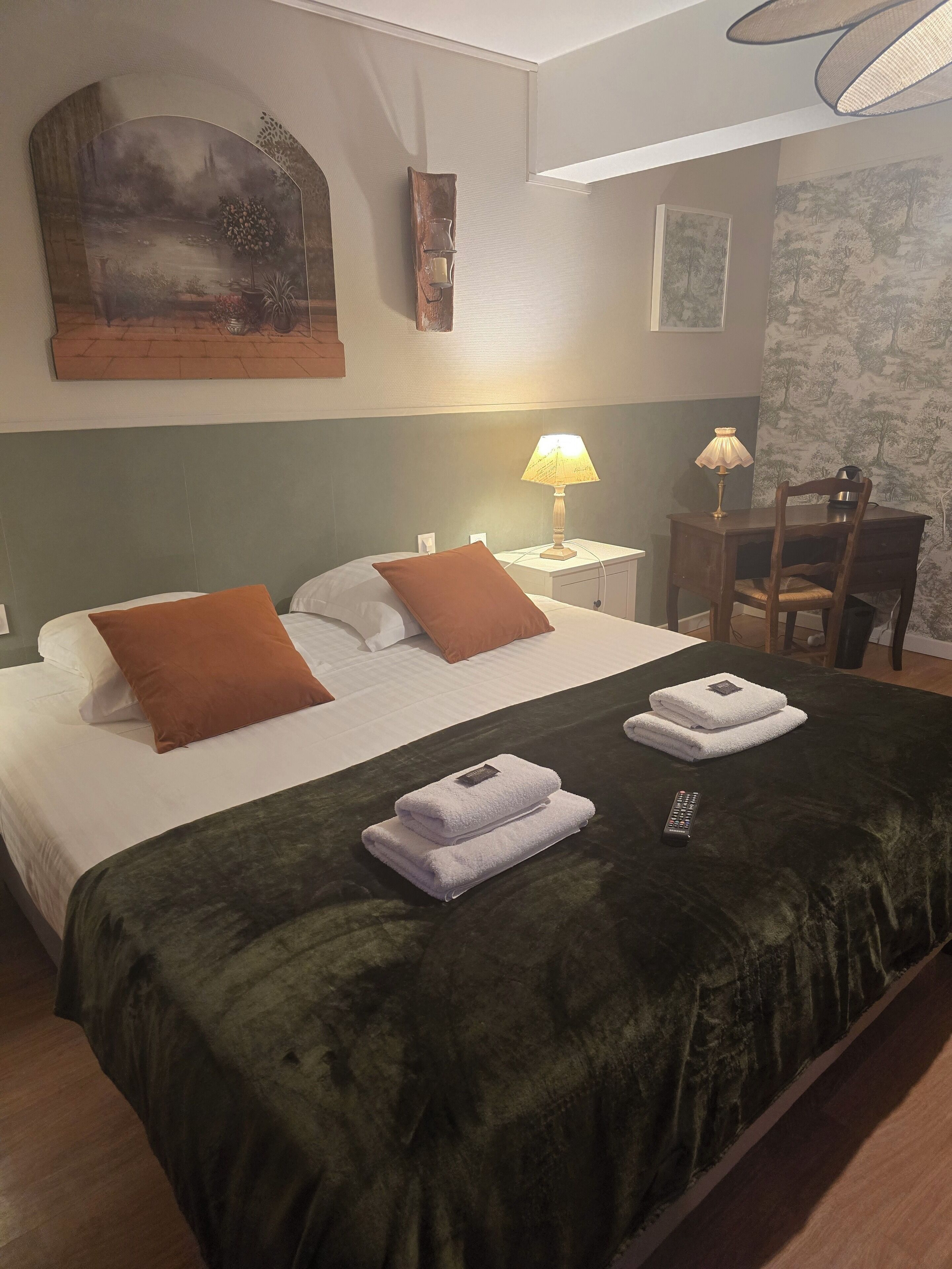 A hotel room with a bed, two bedside lamps, a desk with a chair, and a painting on the wall.