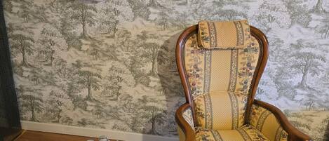 Une pièce dont les murs sont recouverts d’un papier peint à motifs, avec une chaise en bois à l’assise fleurie et une petite table blanche sur laquelle sont disposés divers objets.