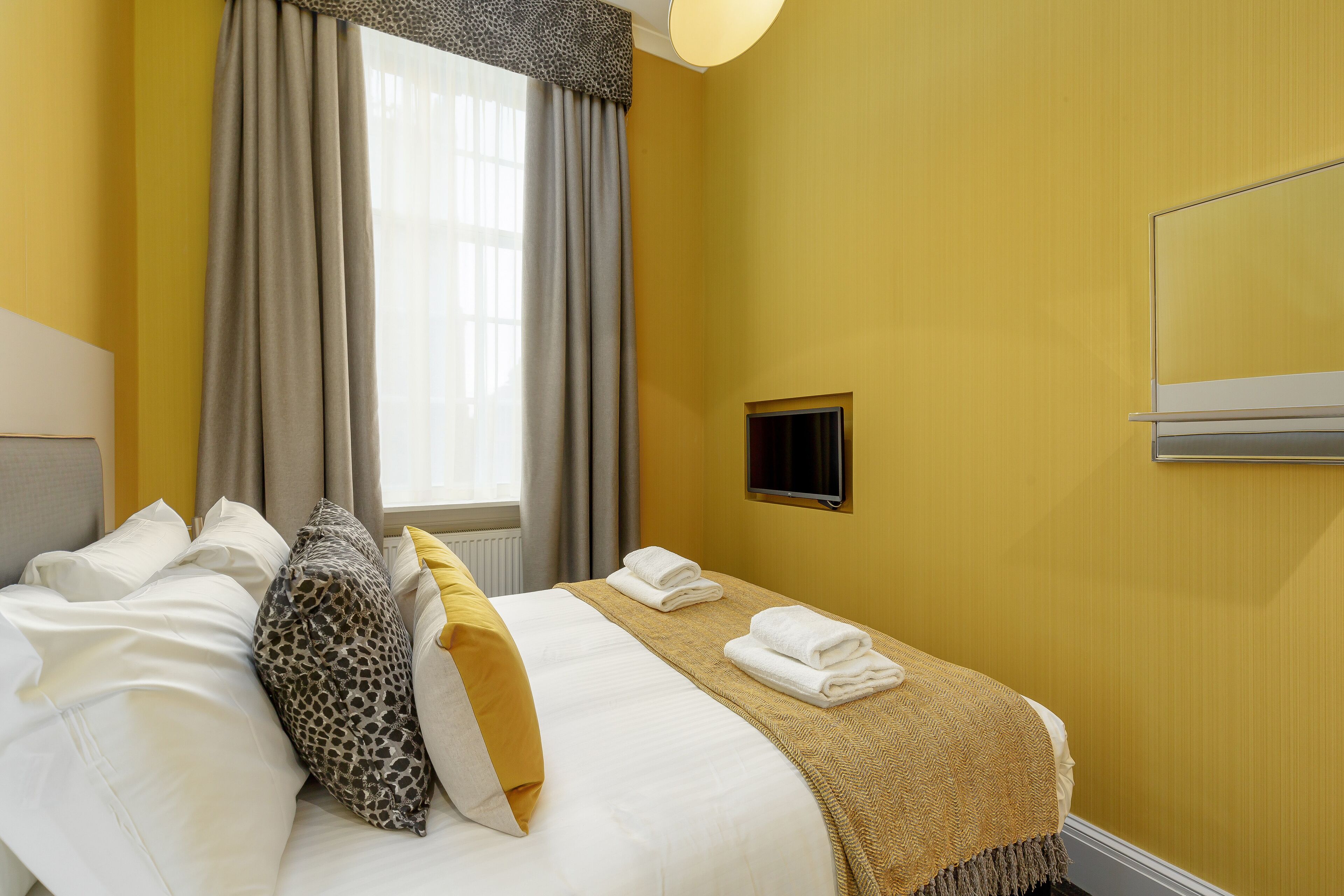 Una habitación de hotel con cama, almohadas, una ventana con cortinas y un televisor en la pared.