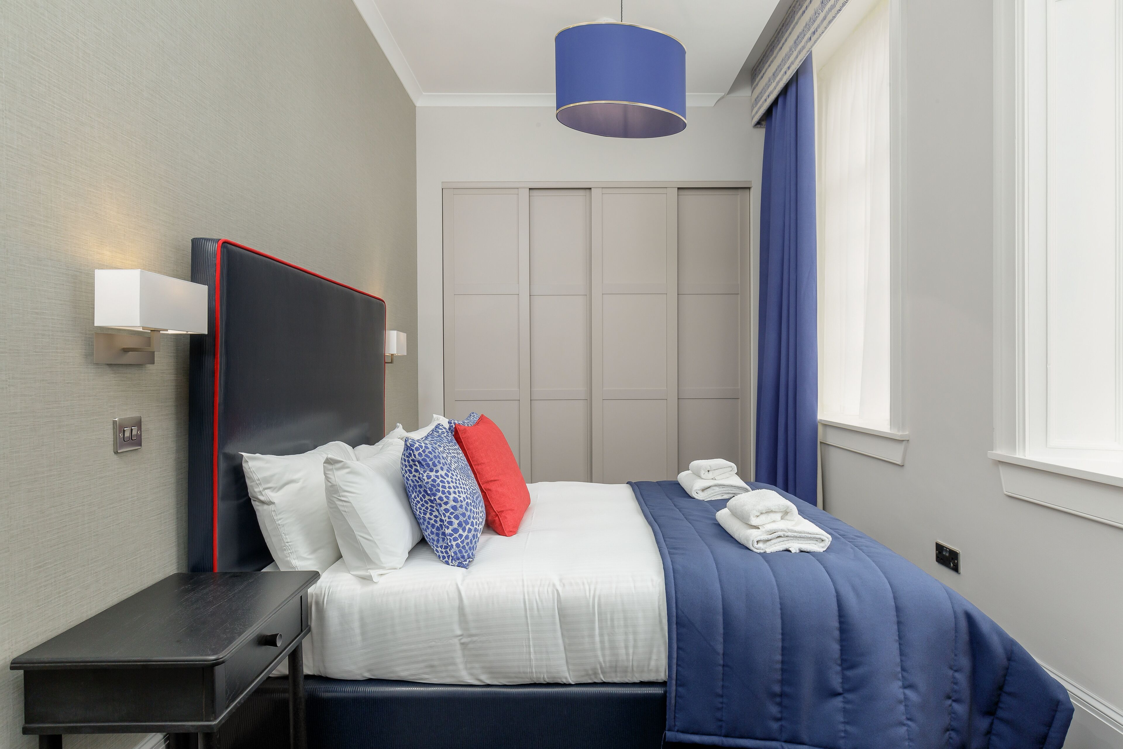 A neatly made bed with white and blue bedding, a red pillow, and a blue blanket. There is a bedside table with a lamp, a window with blue curtains, and a door with a light switch.