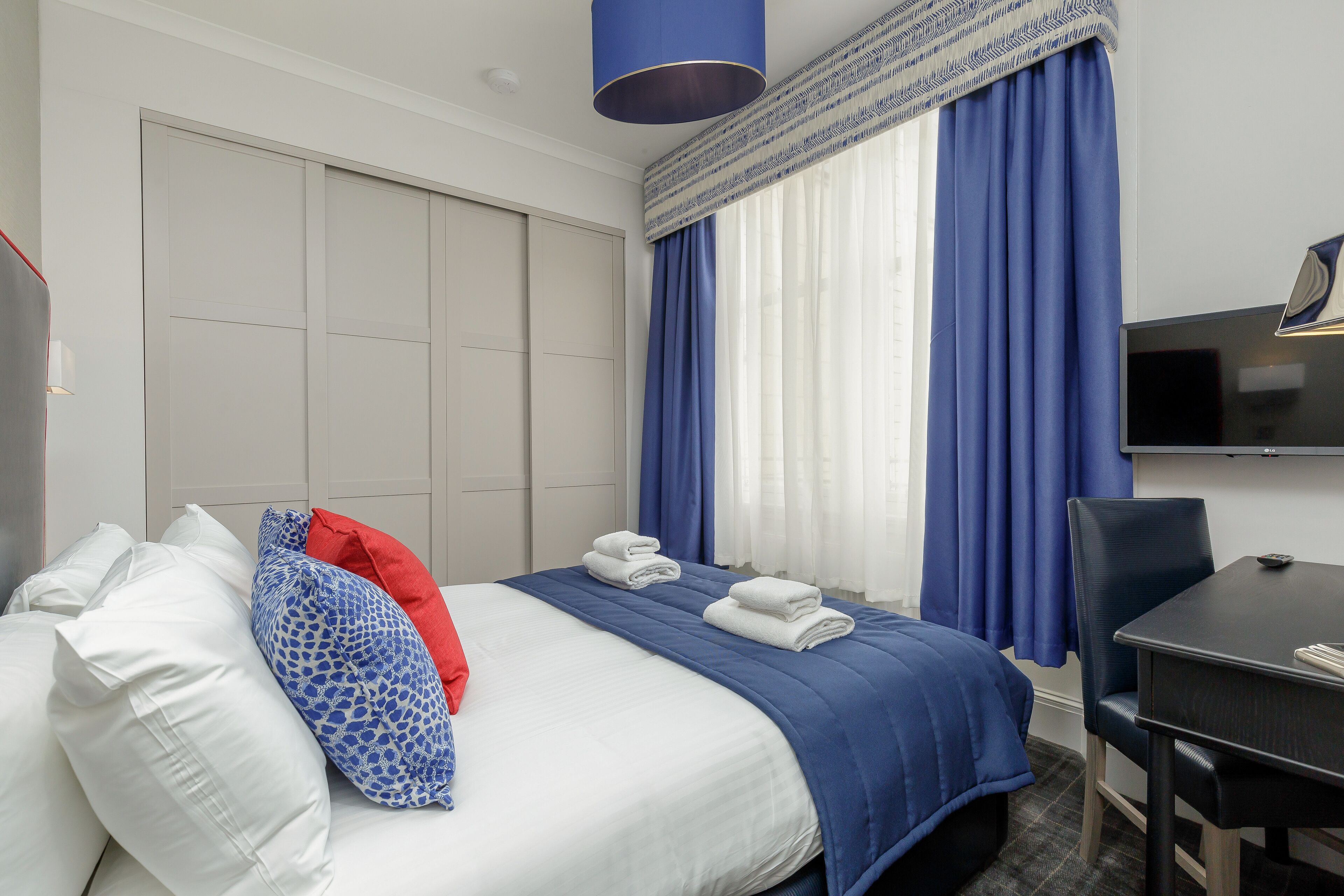 A hotel room with a bed, pillows, a desk, a chair, a television, and a window with curtains.