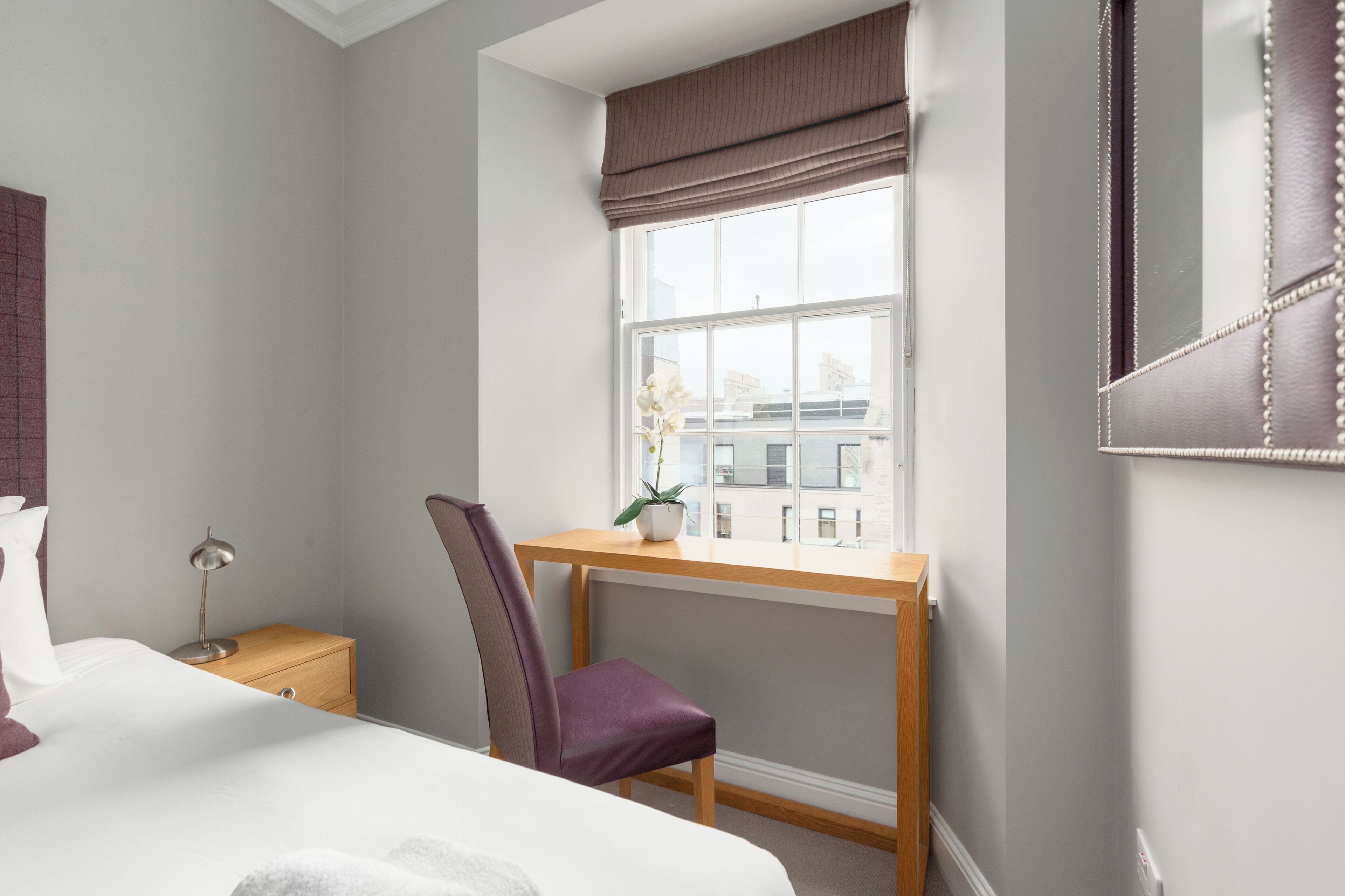 A bedroom with a bed, a desk, a chair, a nightstand, and a window with a view of buildings.