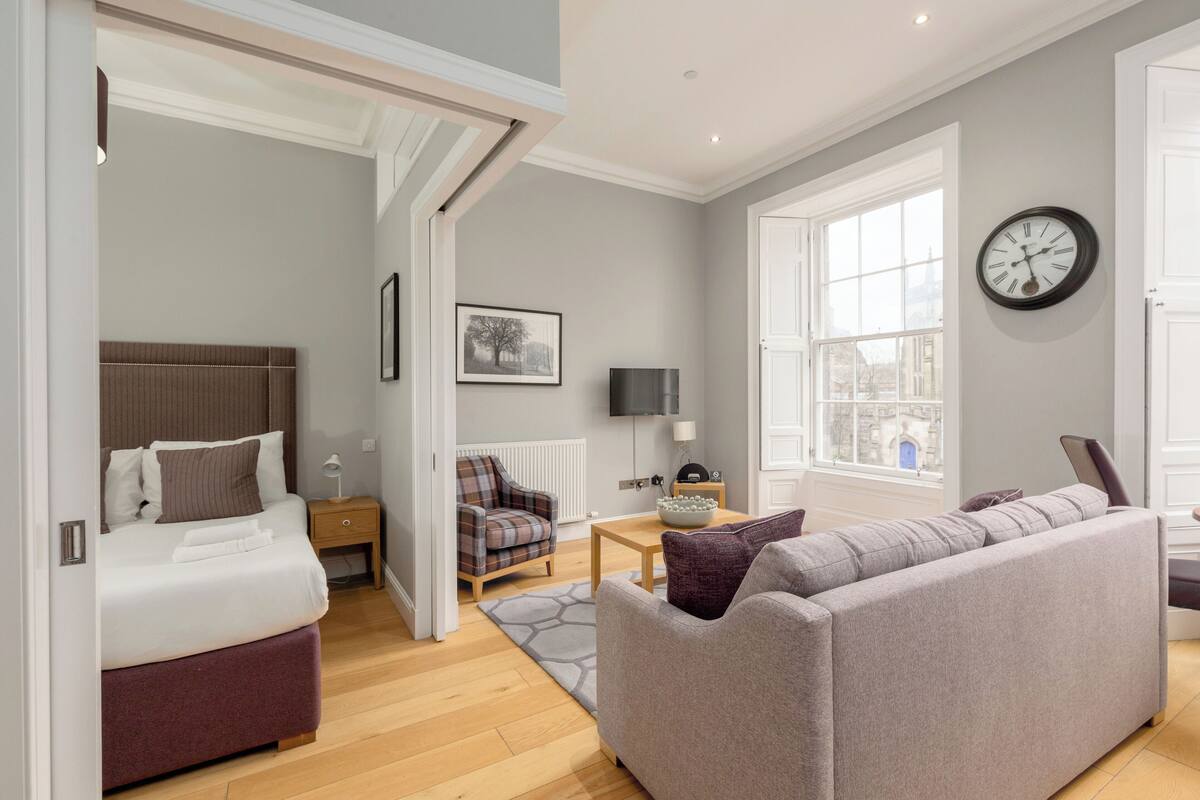 A modern bedroom with a bed, a sofa, and a chair. There is a window with a view of a building outside.