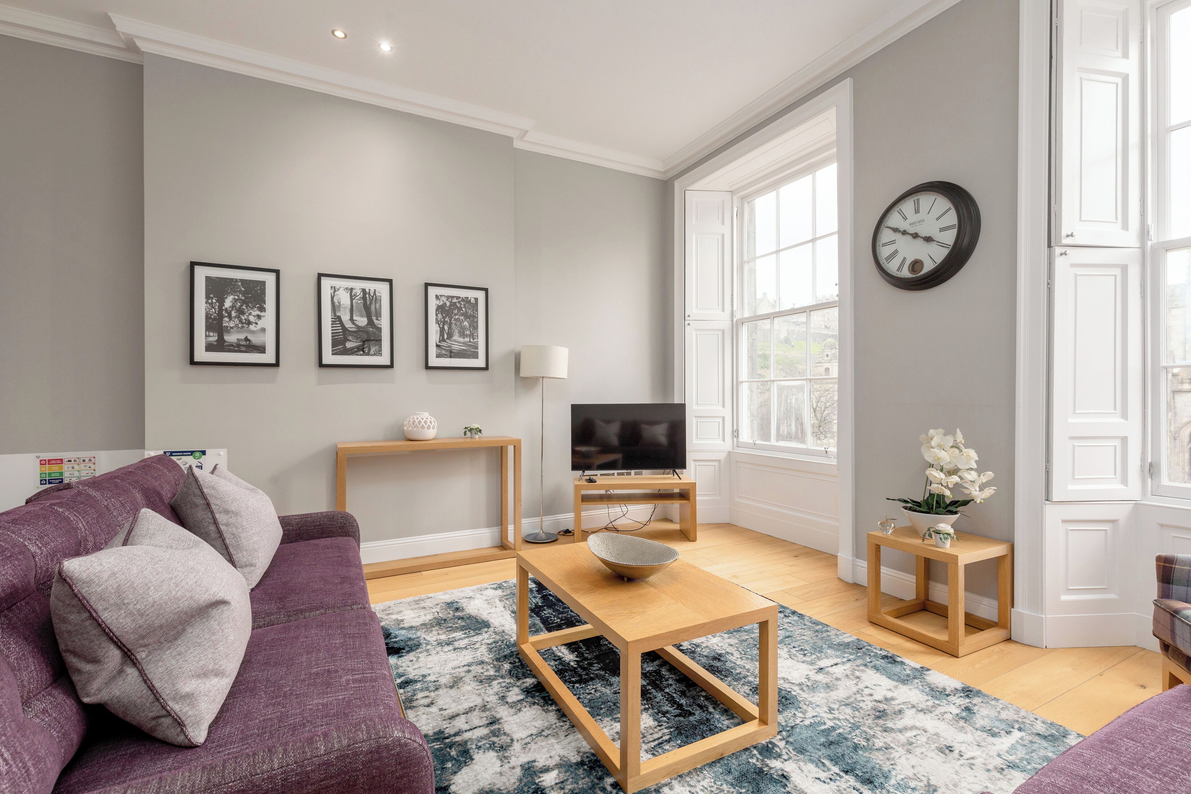 A living room with a purple sofa, a wooden coffee table, a TV on a stand, framed pictures on the wall, and a clock.