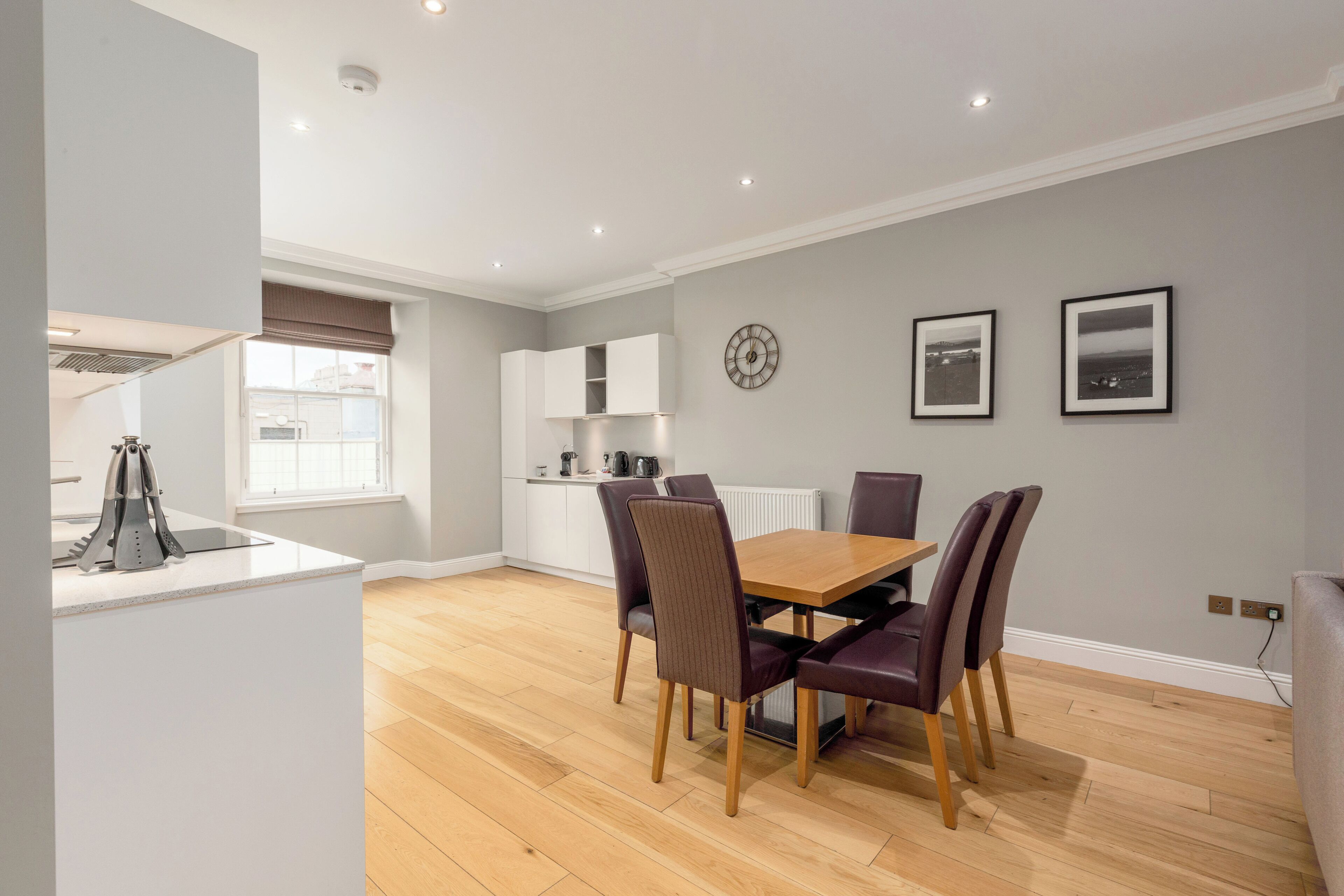 A modern kitchen with white cabinets, a wooden dining table with four chairs, and a window with blinds.