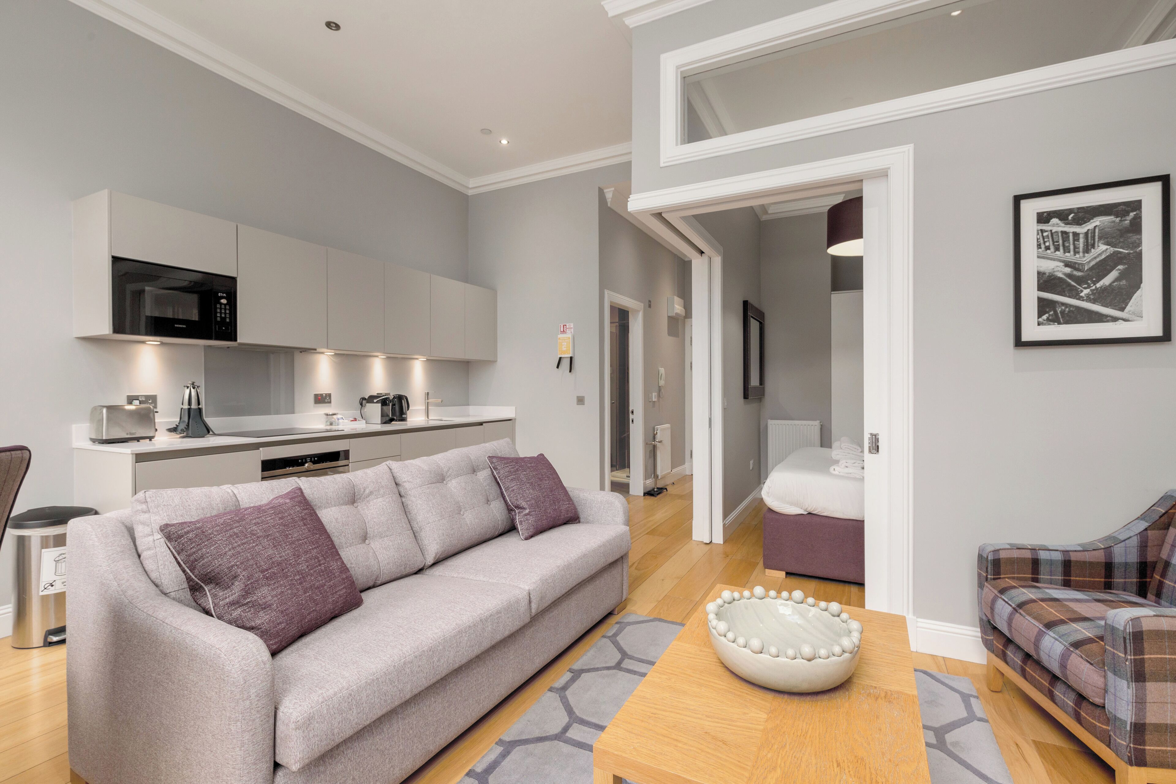 Een moderne woonkamer met een grijze bank, een geruit bankje, een houten salontafel en een keuken met witte kasten en een magnetron.