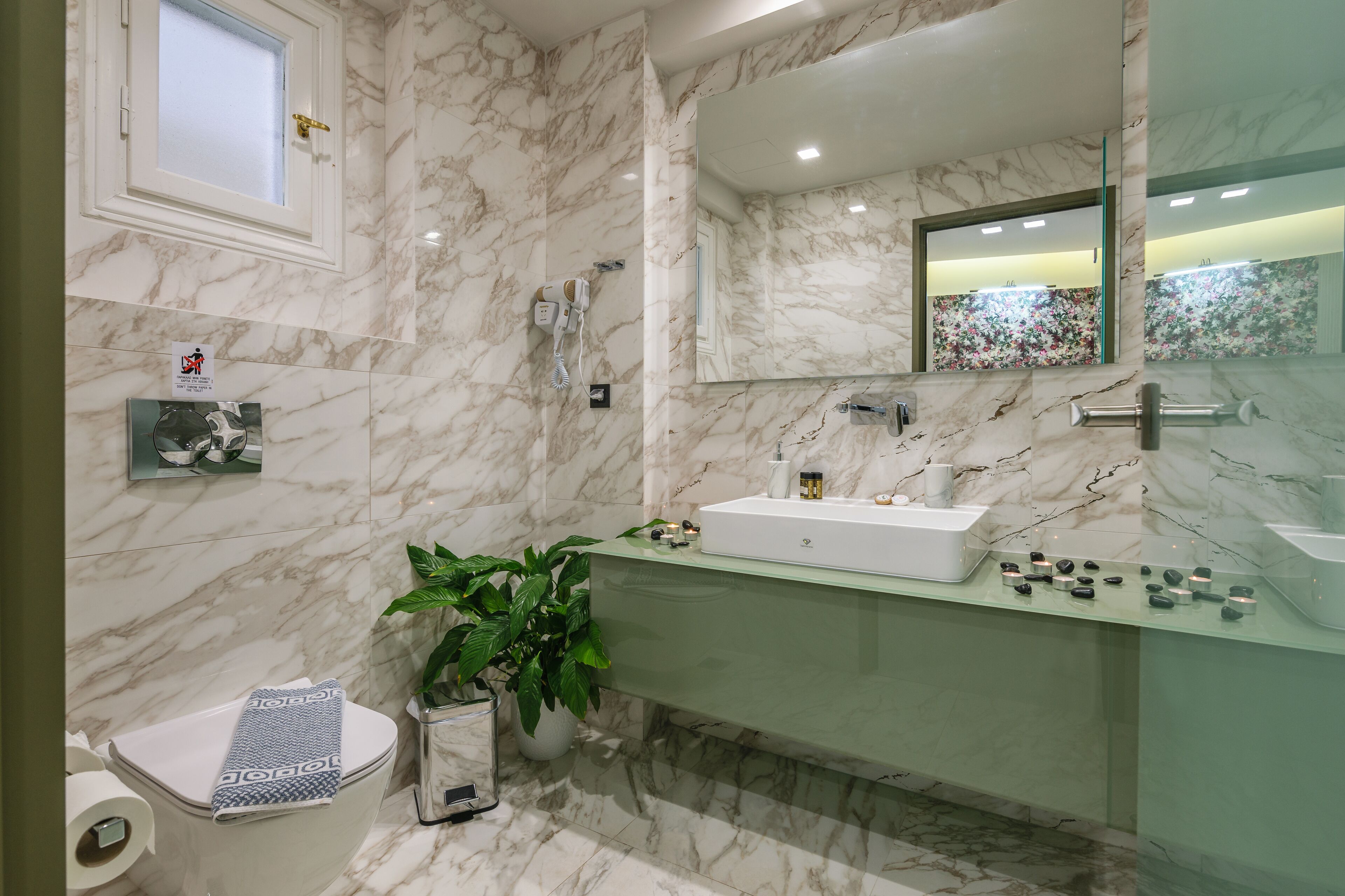 A modern bathroom with a marble floor, a glass sink, a large mirror, and a toilet with a towel and toilet paper.