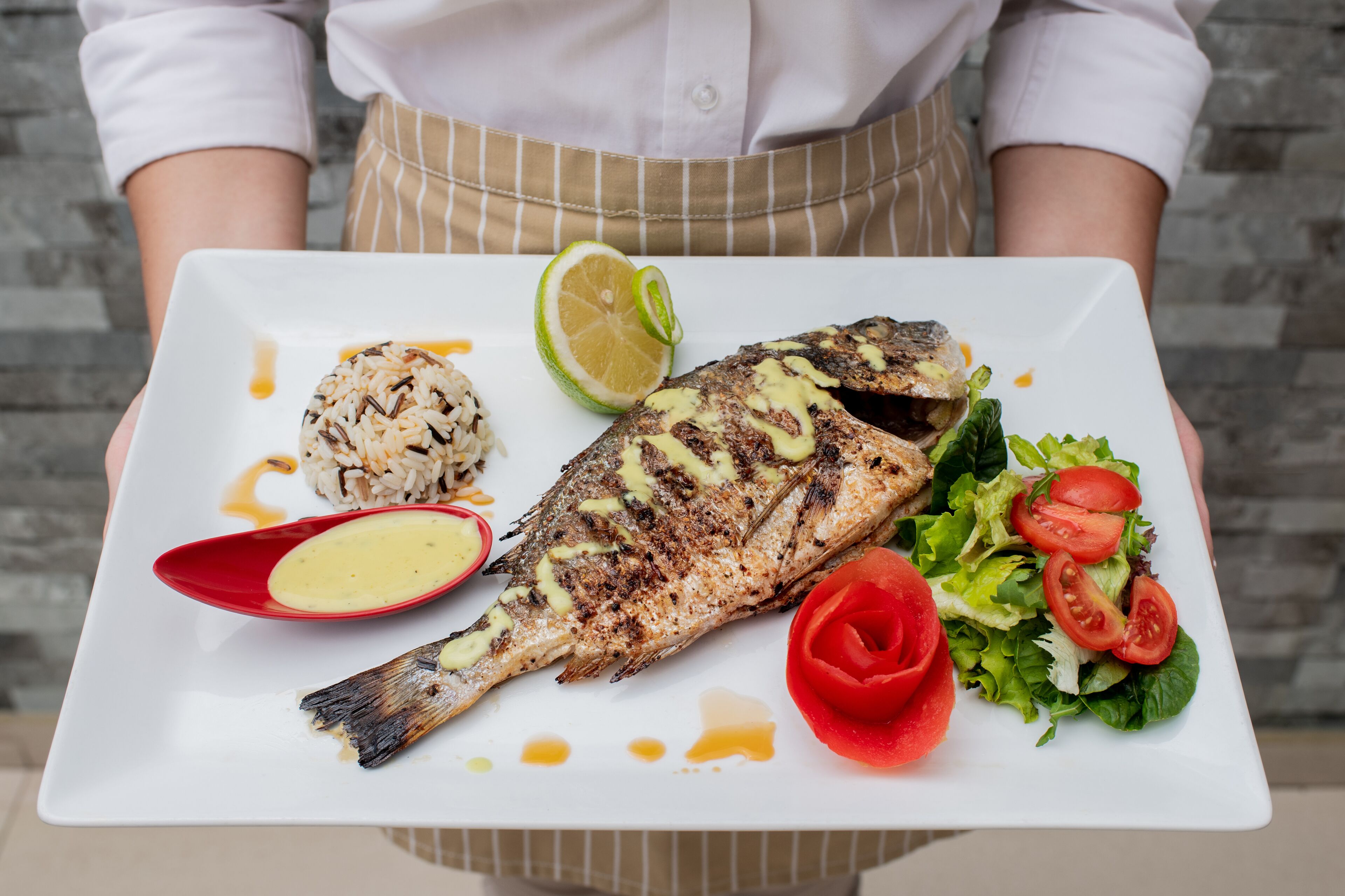 A person holding a white plate with grilled fish, rice, salad, and a slice of lime.