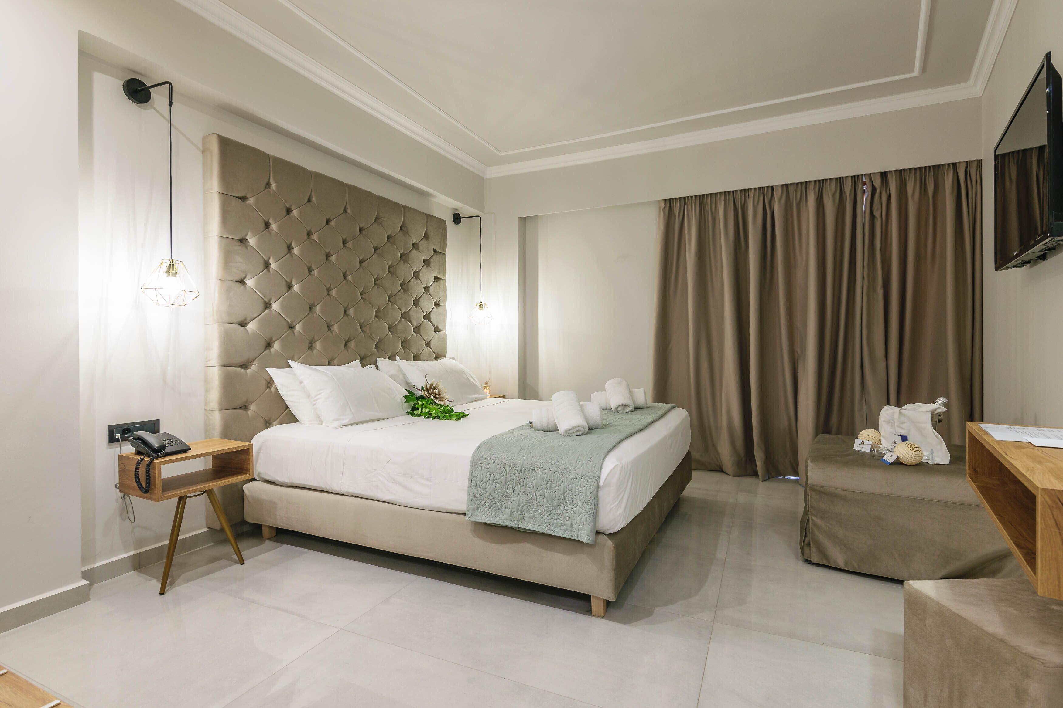 A modern hotel room with a large bed, a bedside table with a phone, a small wooden side table, a TV mounted on the wall, and a window with curtains.