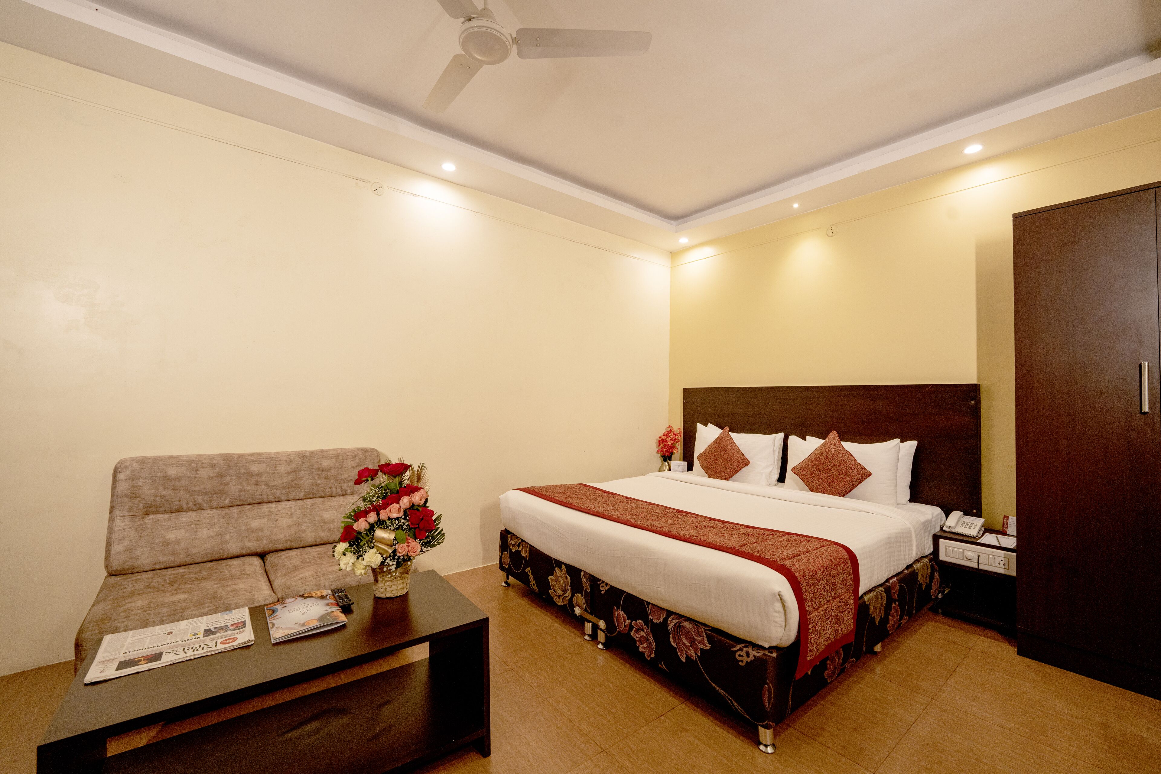 A hotel room with a bed, a sofa, a coffee table with a vase of flowers, and a ceiling fan.