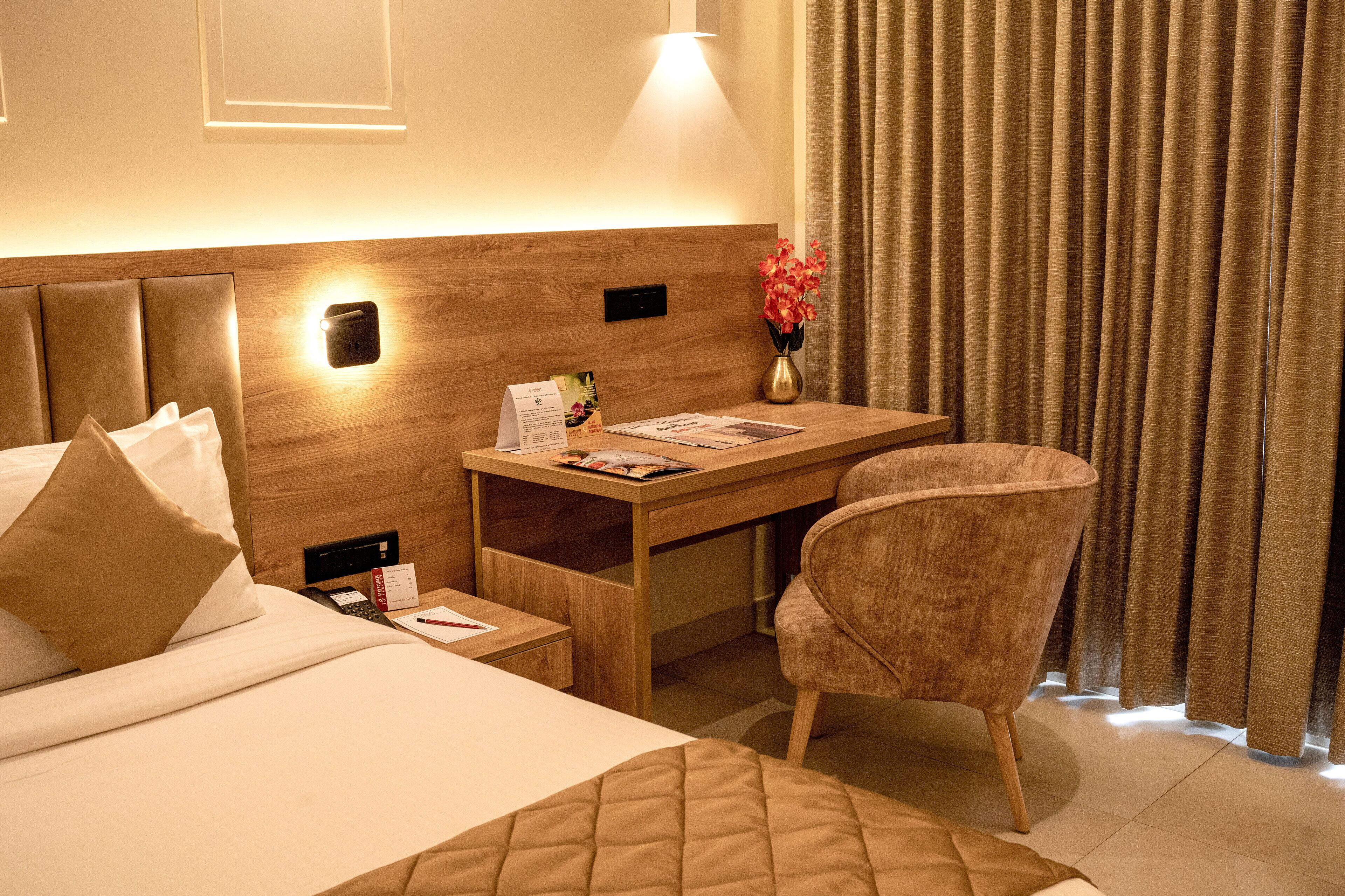 Habitación de hotel con cama, escritorio, silla, mesita de noche, lámpara, un florero con flores y una cortina.