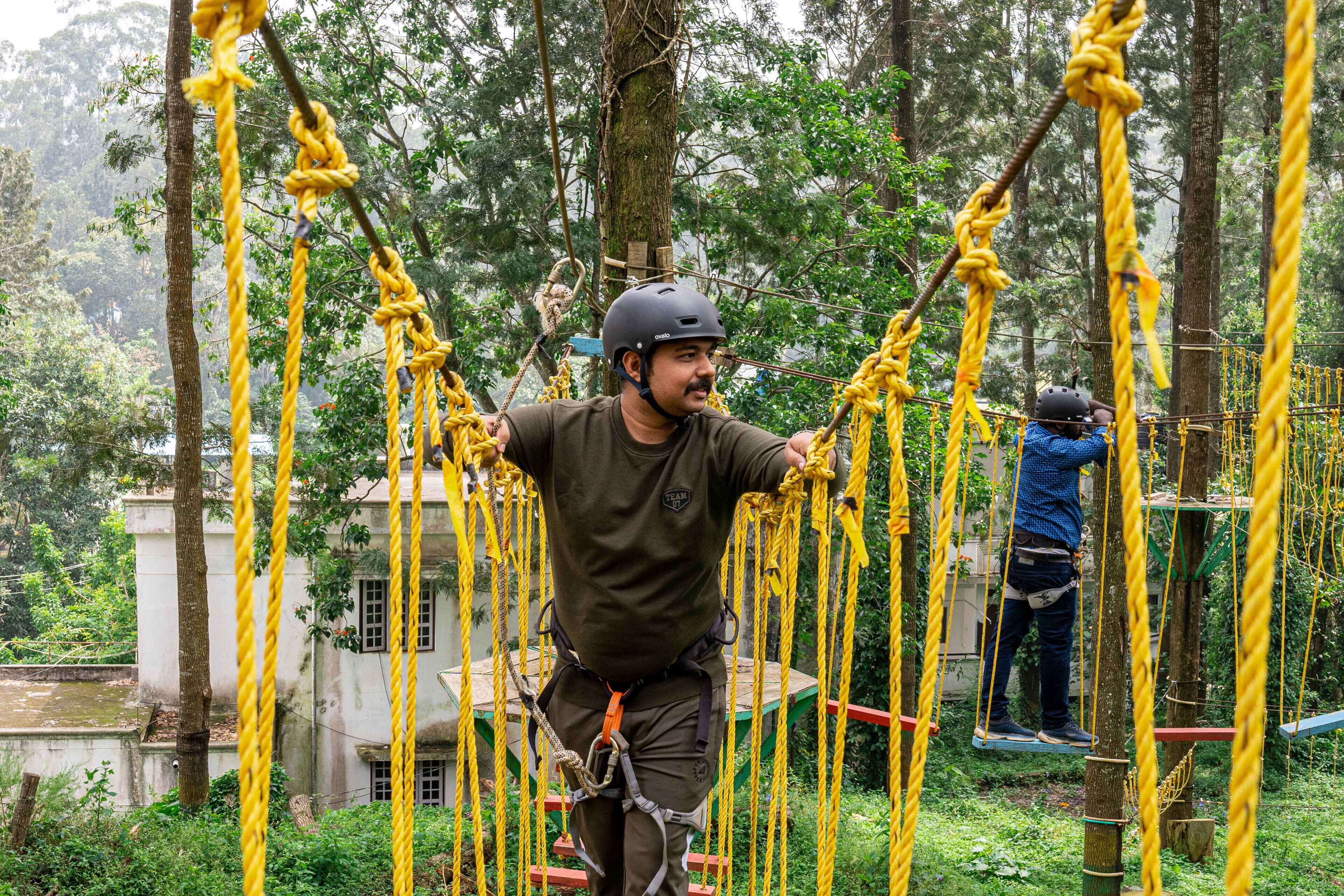 A person navigating a rope course in a forested area with trees and a building in the background.