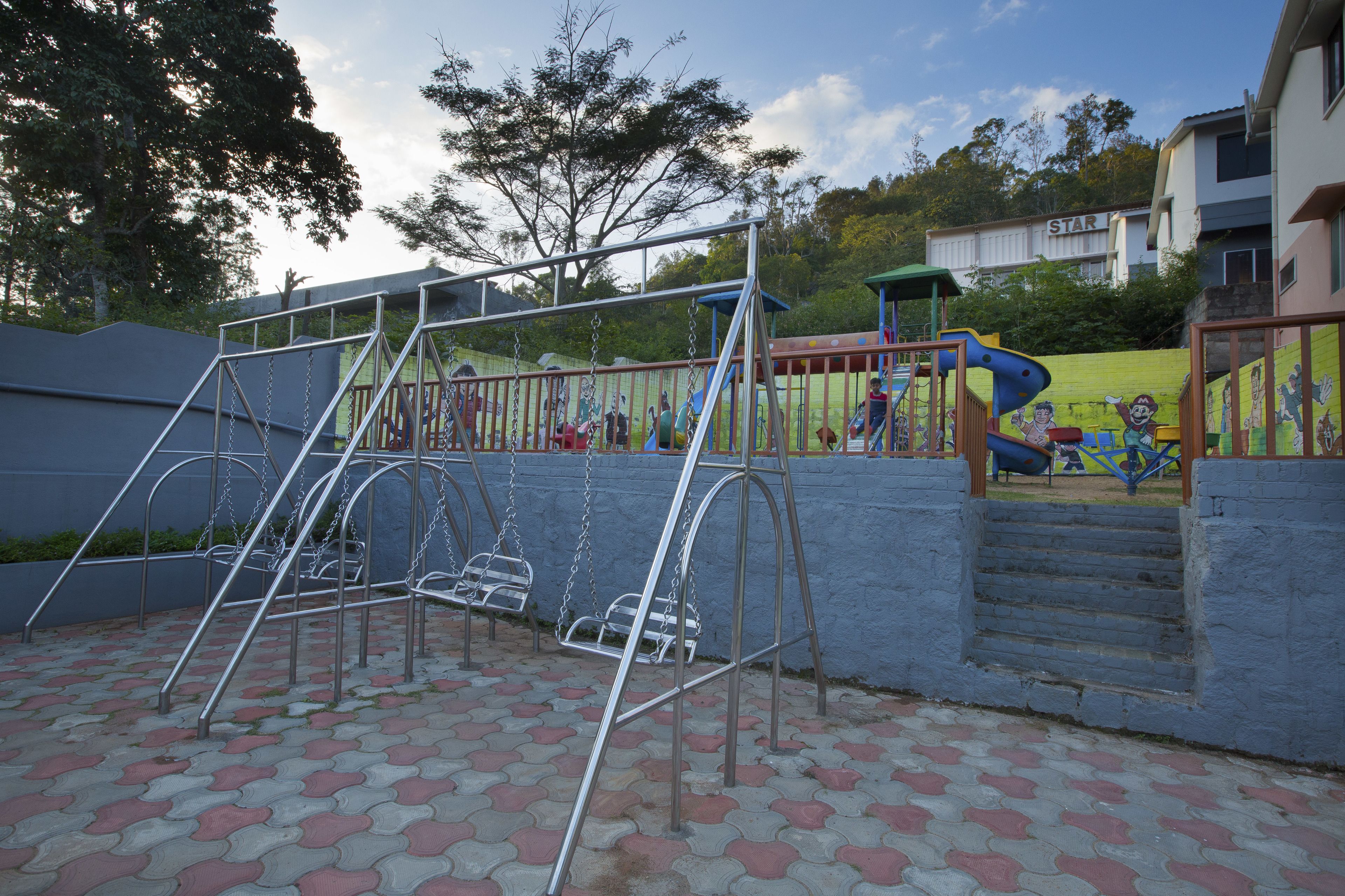 Un parc infantil amb cadira d'oscil·lació i tobogan, envoltat per una paret de maó i un edifici amb la inscripció “STAR” al fons.