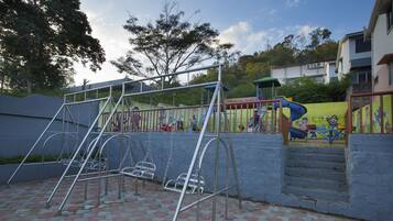 Un parc infantil amb cadira d'oscil·lació i tobogan, envoltat per una paret de maó i un edifici amb la inscripció “STAR” al fons.