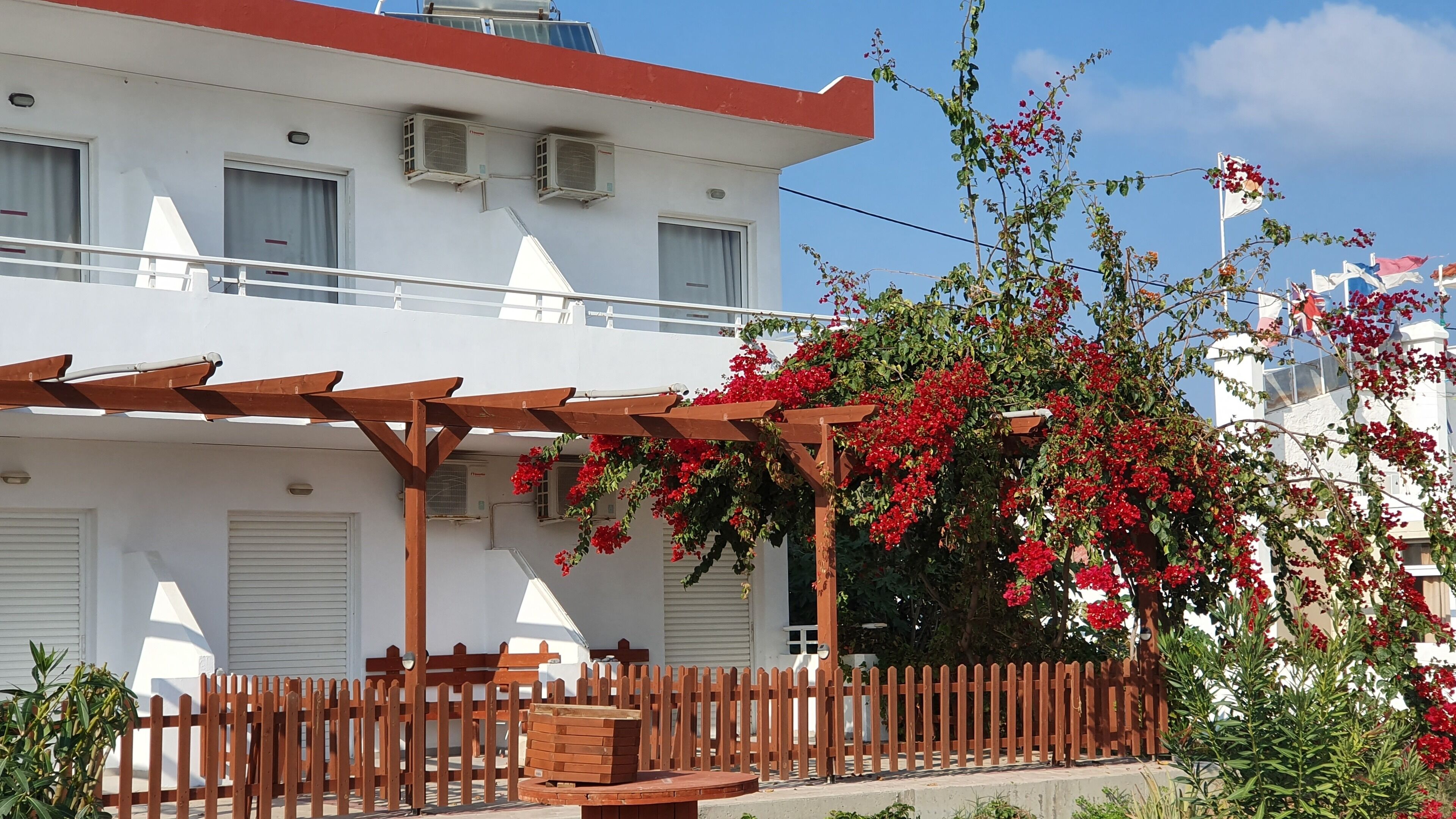 A white building with a red roof, air conditioning units, and a wooden pergola.
