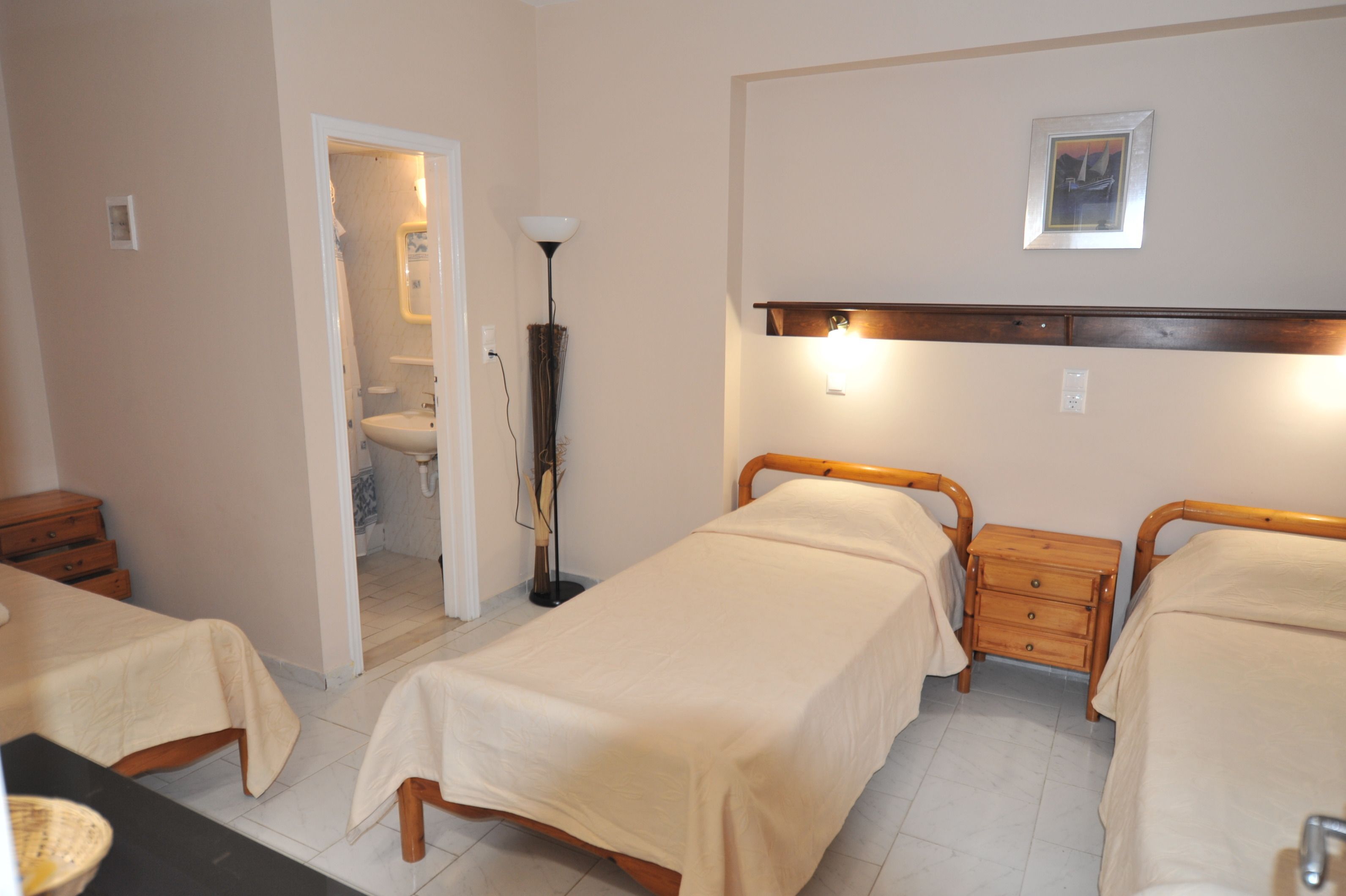 Une chambre d’hôtel avec deux lits simples, une table de chevet avec des tiroirs, une petite table, une lampe, un tableau encadré au mur et une porte ouverte donnant sur une salle de bain.
