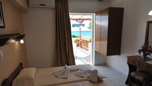 Habitación de hotel con dos camas, un escritorio con silla, una mesita con una taza y un control remoto, y vista a la playa a través de una puerta abierta.