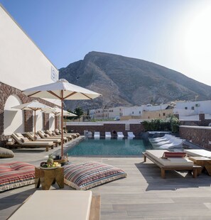 A poolside area with lounge chairs and umbrellas, a stone wall, and a mountain in the background. - Selini Hotel (Santorini)