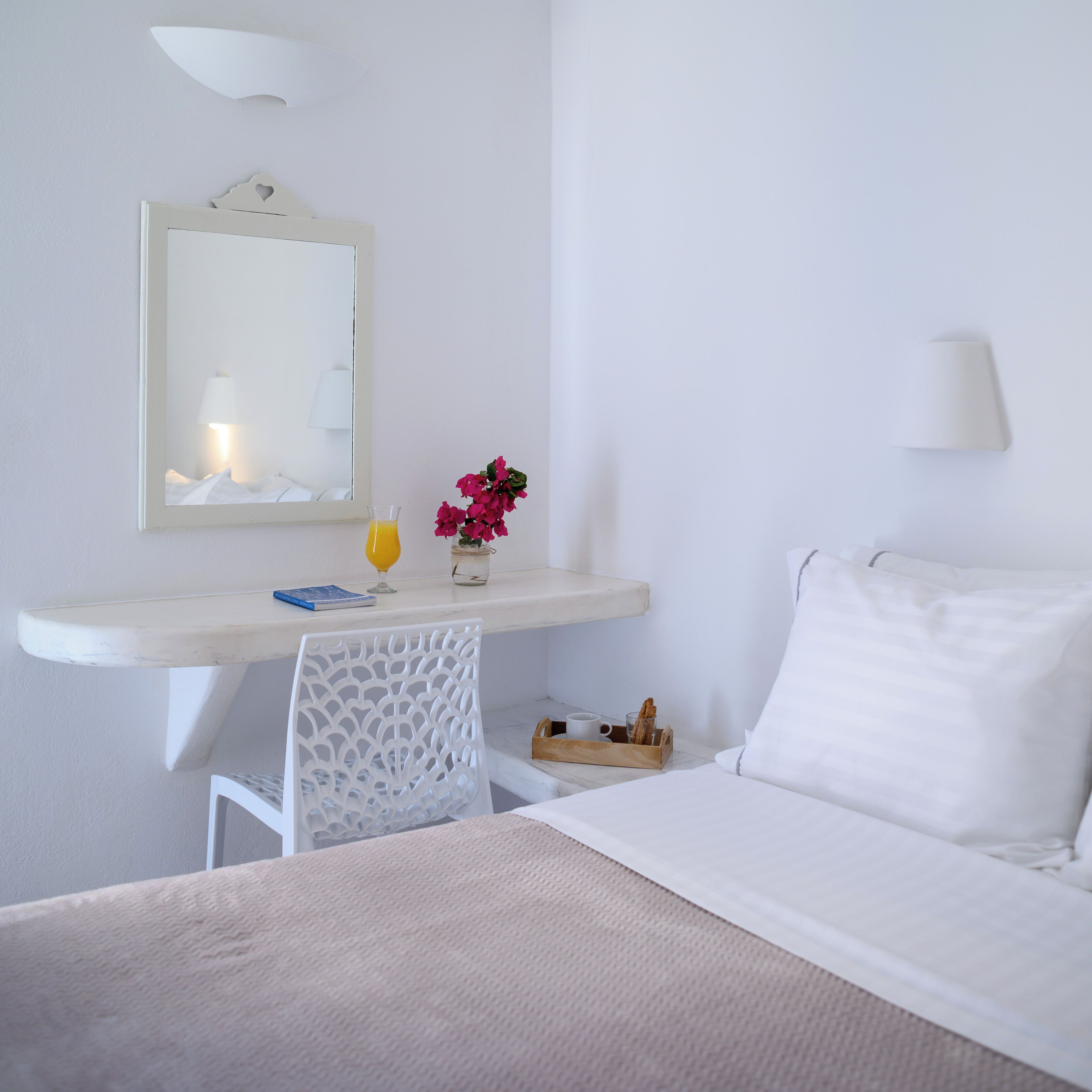 A hotel room with a bed, a desk with a chair, a mirror, a vase with flowers, a glass of orange juice, and a small table with bread.