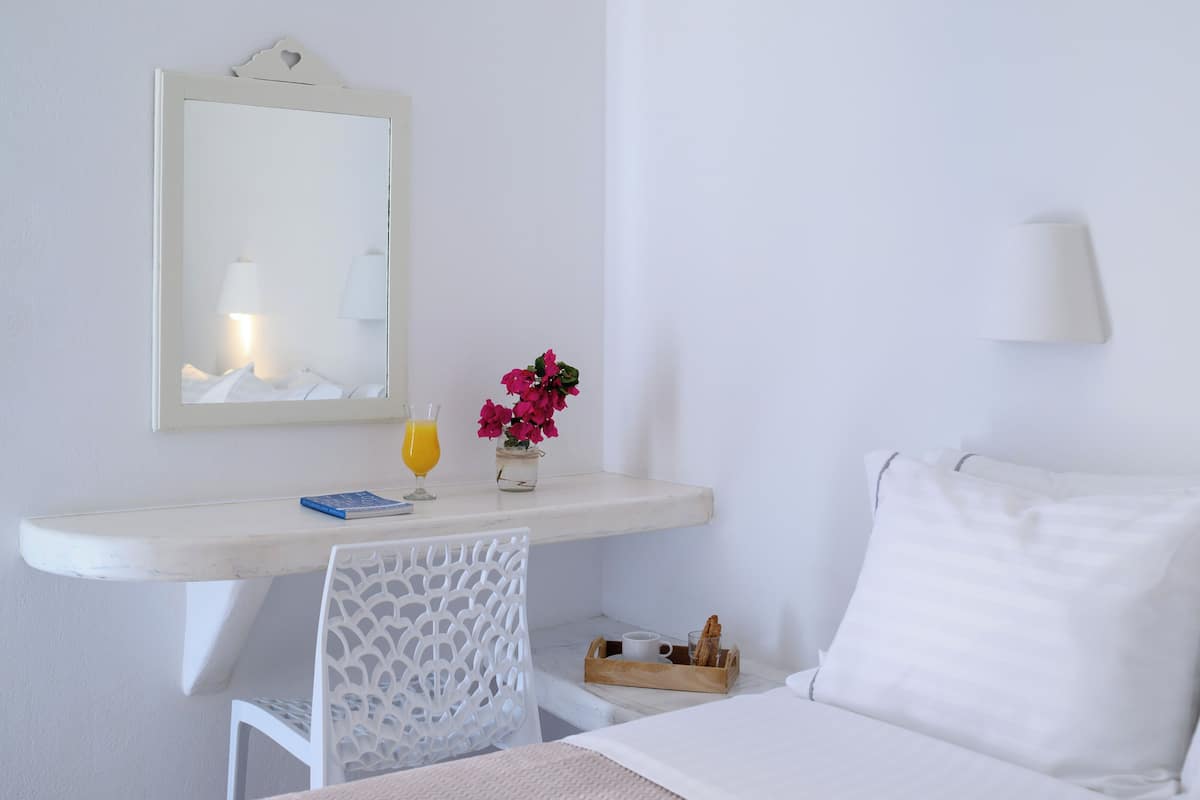 A hotel room with a bed, a desk with a chair, a mirror, a vase with flowers, a glass of orange juice, and a small table with bread.