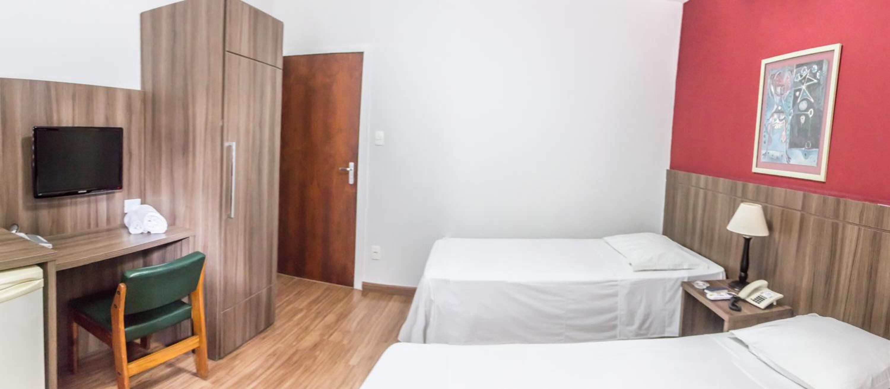 Quarto de hotel com guarda-roupa de madeira, uma escrivaninha com cadeira, cama, mesa de cabeceira com abajur e televisão na parede.